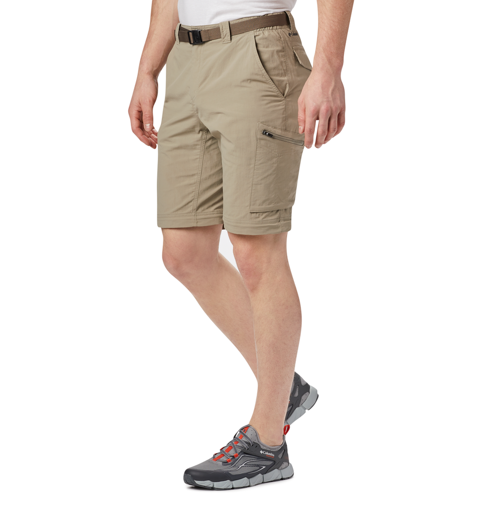 אדם לבוש במכנסי קולומביה IL Silver Ridge II Convertible עם כיס רוכסן, חגורה שחורה ונעלי טיולים אפורות עם נגיעות אדומות עומד על רקע לבן.