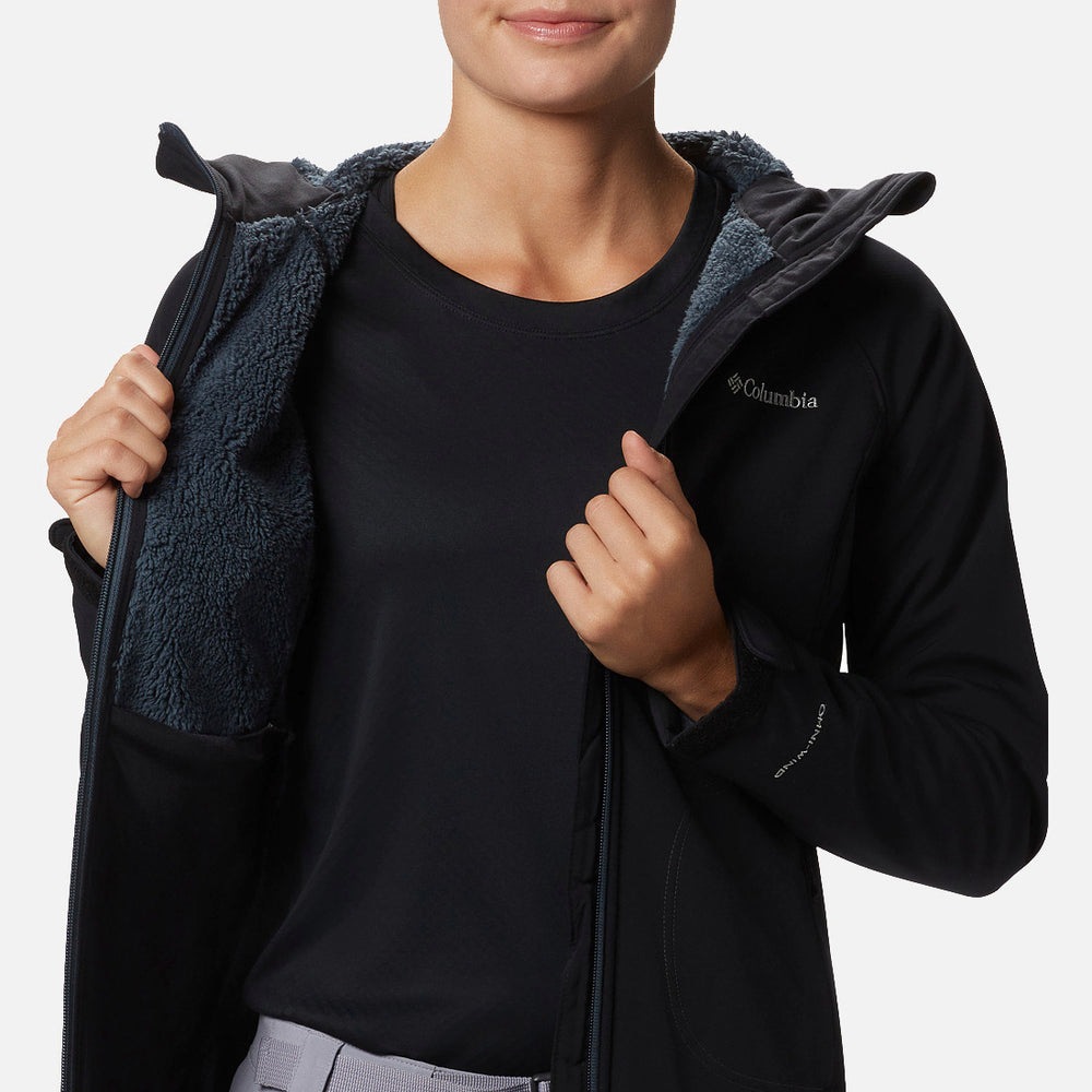 אדם מחזיק פתוחה מעיל Softshell Phurtec II בצבע שחור של המותג ColumbiaIL, עם בטנה אפורה ומרופדת, וחושף את פנים המעיל ואת החולצה השחורה שמתחתיו. הרקע הלבן הפשוט מדגיש את המעיל האופנתי הזה המיועד לפעילויות בחוץ.