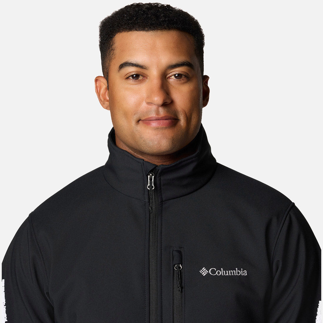 גבר לבוש במעיל Softshell של ColumbiaIL Ascender בצבע שחור עם צווארון גבוה עומד על רקע לבן חלק, פונה למצלמה ומחייך קלות.