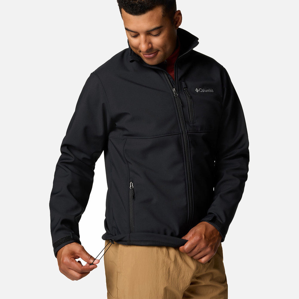 אדם לבוש במעיל Softshell שחור של ColumbiaIL Ascender ובמכנסיים בצבע חום בהיר מכוון את שרוך השוליים, מחייך ומביט מטה, על רקע בהיר וחד-גוני.