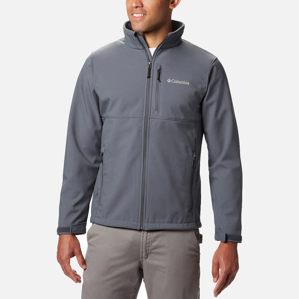 גבר לבוש במעיל רוכסן אפור מדגם Ascender Softshell של ColumbiaIL, בעל צווארון גבוה וכיסים עם רוכסן, ומכנסיים אפורים, עומד על רקע לבן חלק.