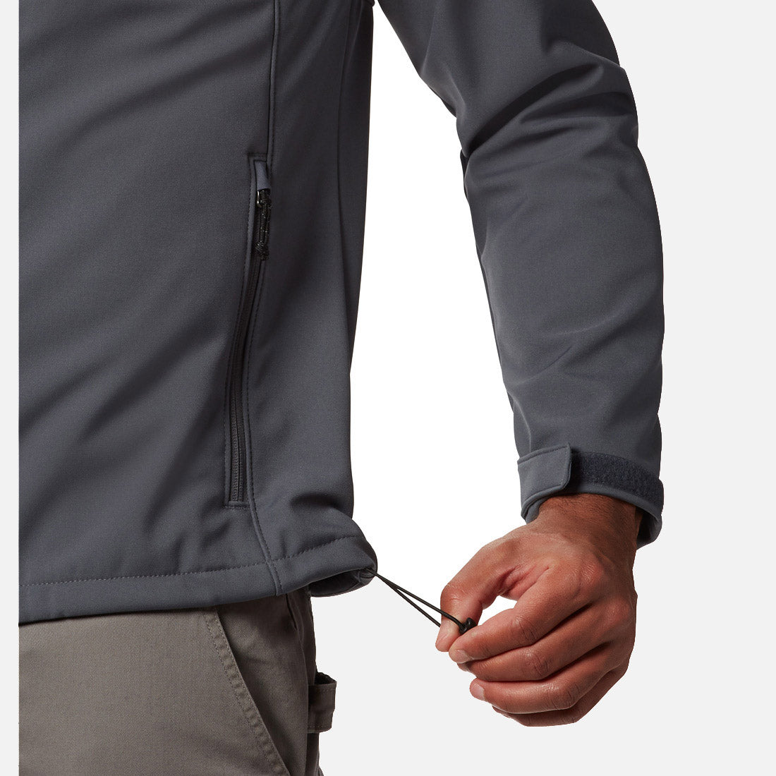 אדם מהדק את שרוך ההידוק בשולי מעיל ה-Softshell האפור מדגם Ascender של ColumbiaIL, הכולל כיס עם רוכסן ורצועת וולקרו בשורש כף היד, בשילוב עם מכנסיים אפורים תואמים.
