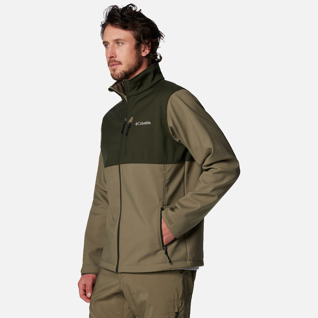 גבר לבוש במעיל Softshell של ColumbiaIL Ascender בצבעי זית וירוק כהה, עם כיסים עם רוכסן ומכנסיים תואמים, עומד כשידו האחת בכיס, מביט לצד על רקע לבן חלק.