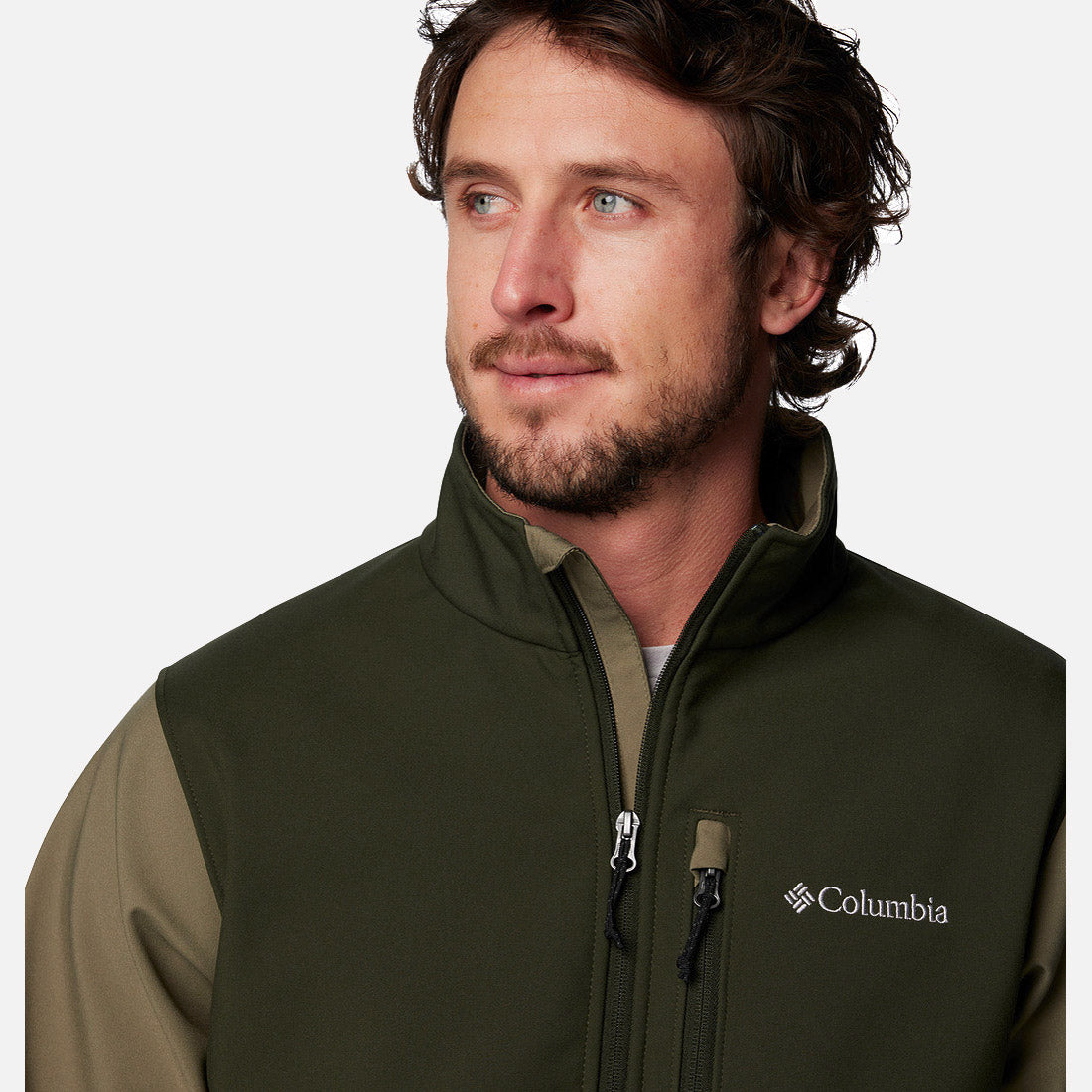 גבר בעל שיער חום גלי וזקן לובש מעיל Softshell בצבע ירוק זית של המותג ColumbiaIL Ascender, ומביט הצידה על רקע לבן חלק.