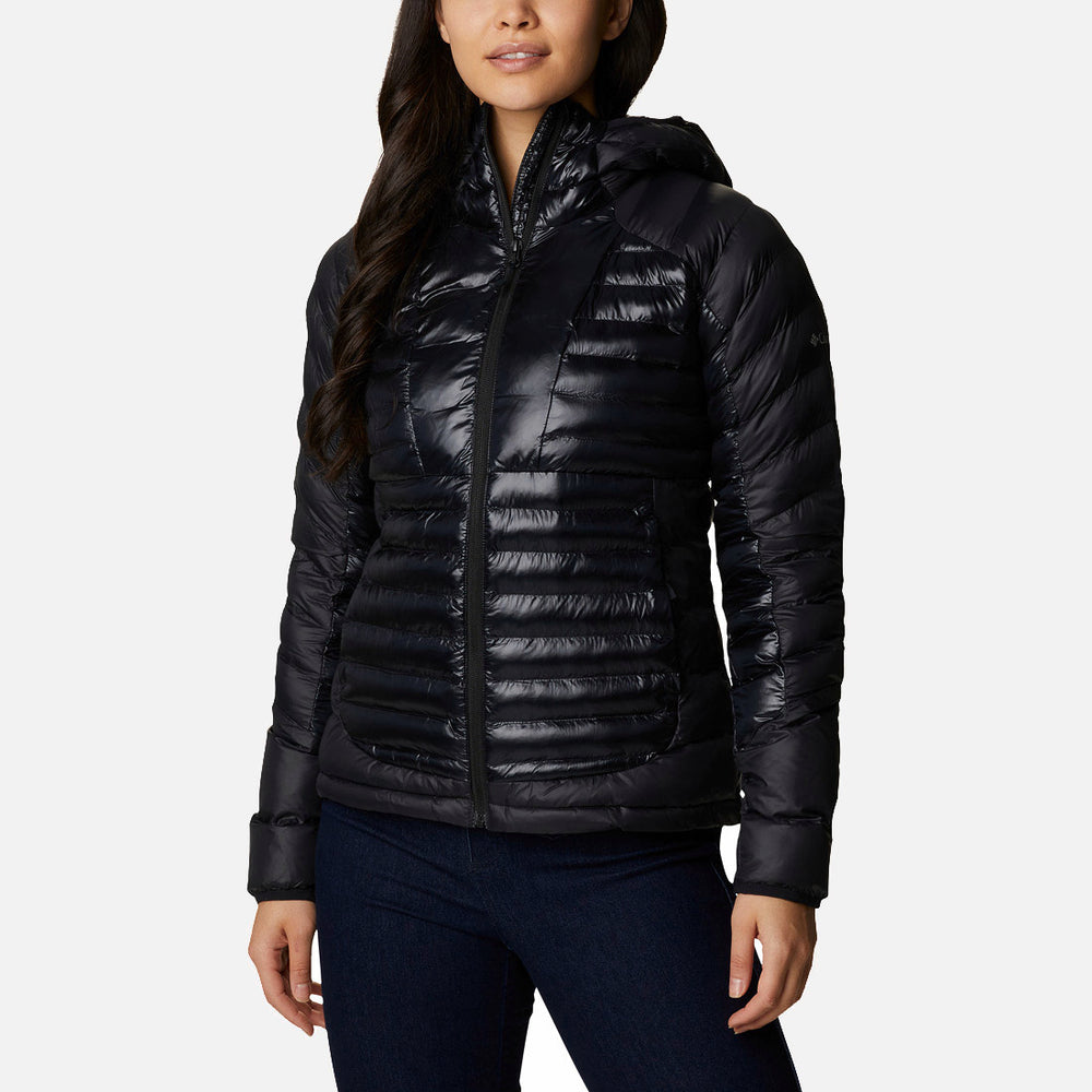 אישה מדגמנת את ColumbiaIL Labyrinth Loop Hooded W, מעיל פוך שחור מבריק עם ברדס, בשילוב עם ג'ינס כהה ונעלי ספורט. היא עומדת על רקע לבן, ושערה הארוך והכהה משלים את המראה האופנתי.