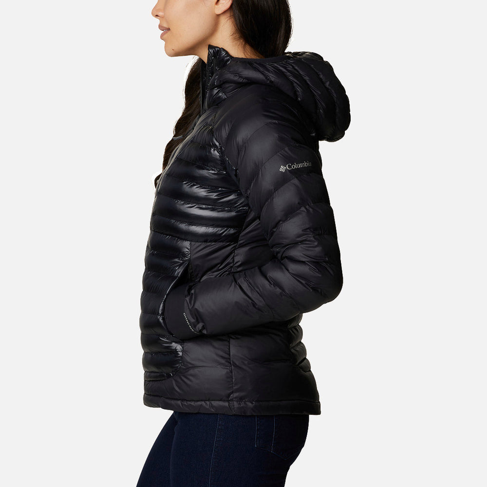 אישה לבושה במעיל פוך שחור עם ברדס מדגם "ColumbiaIL Labyrinth Loop Hooded W" עומדת בפרופיל, ידיה בכיסים, ומשלימה את המראה עם נעלי ספורט מסוגננות מדגם "LABYRINTH LOOP" על רקע לבן חלק.
