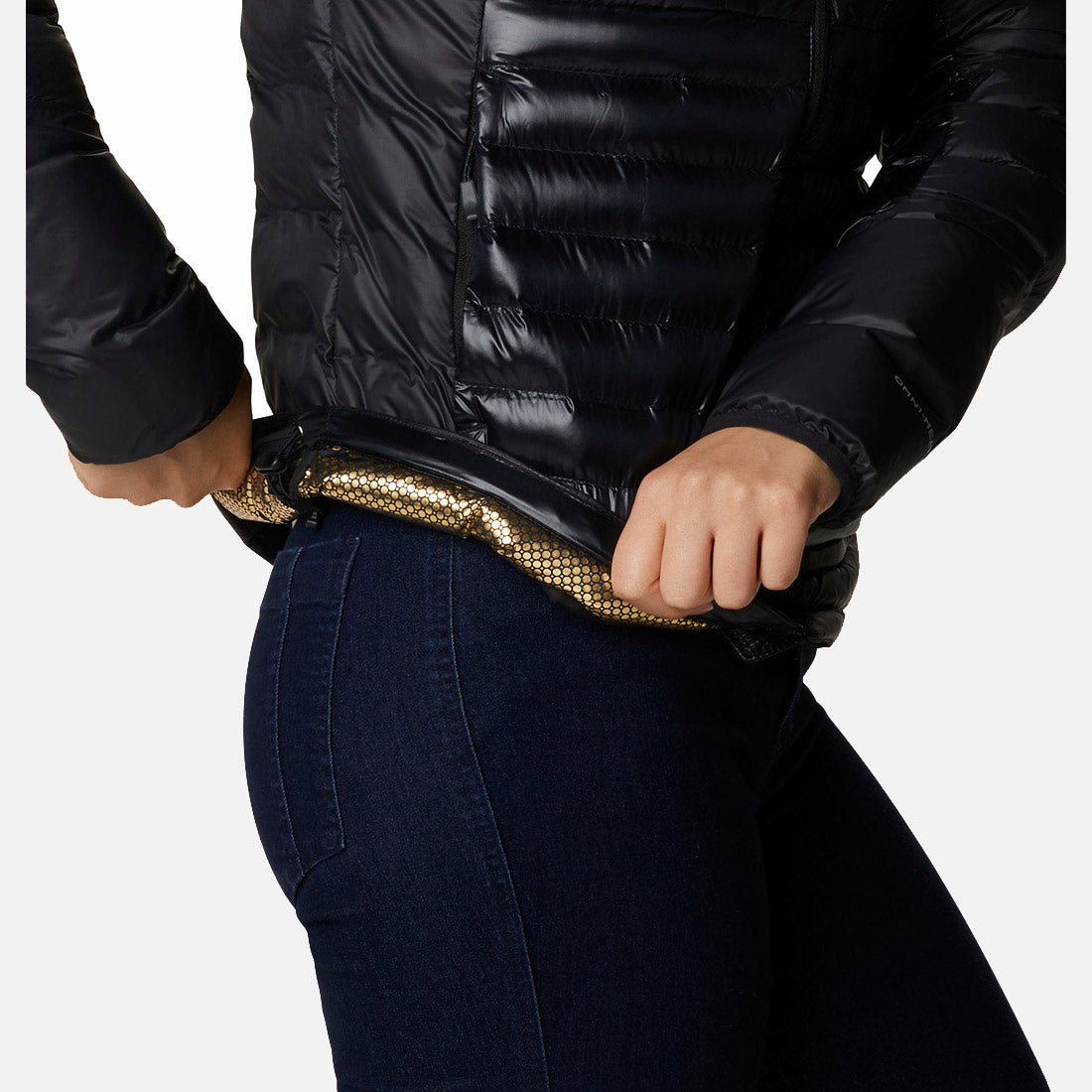 אדם לבוש במעיל פוך שחור ומבריק מדגם "Columbia IL Labyrinth Loop Hooded W" פותח את הרוכסן וחושף בטנה זהובה בדוגמת חלת דבש, בשילוב עם ג'ינס כחול כהה ונעלי ספורט אלגנטיות.