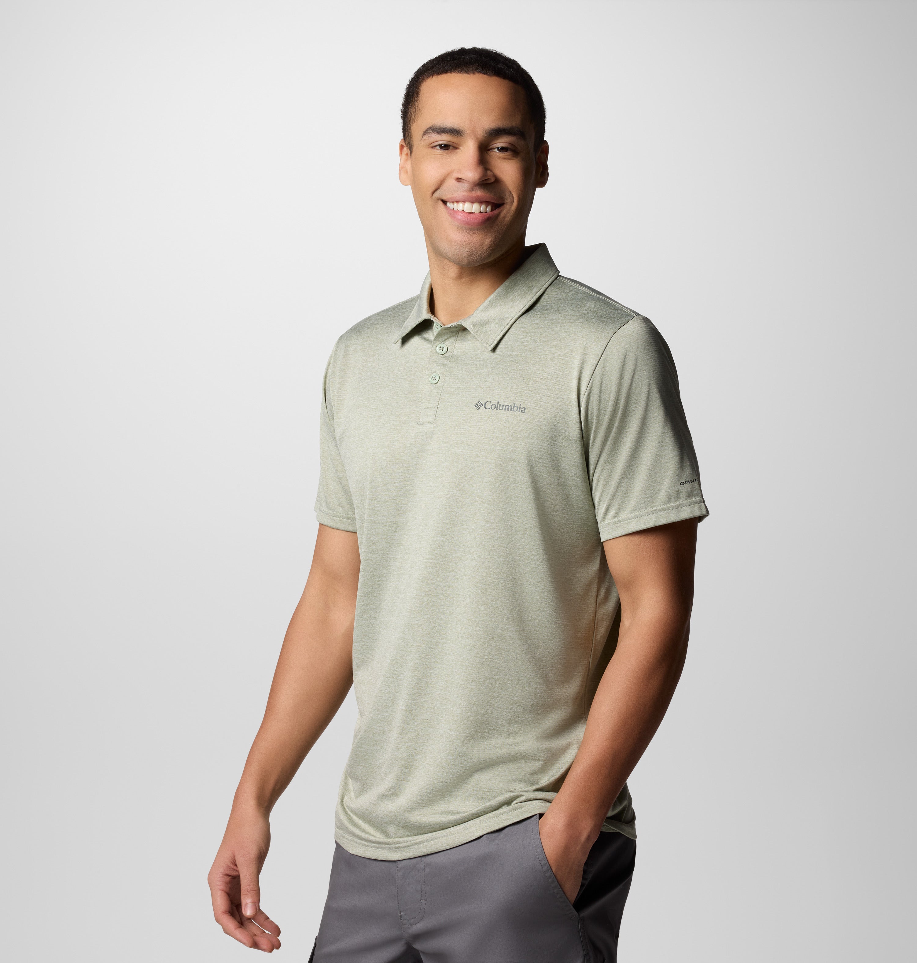 גבר מחייך לובש את חולצת הפולו "Columbia Hike Polo" מבית קולומביה ישראל בצבע ירוק בהיר, עם מכנסיים קצרים אפורים, עומד על רקע בהיר ופשוט, כשידו האחת בכיס.
