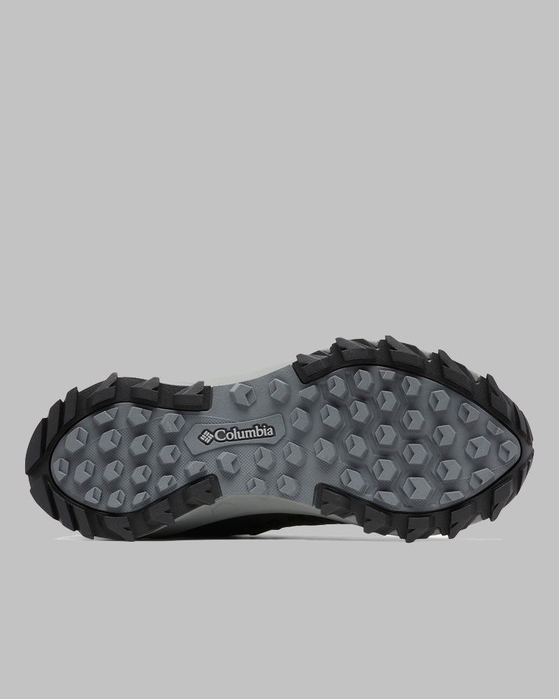 בתמונה נראית הסוליה של נעל הנשים ColumbiaIL Peakfreak II Outdry W, הכוללת פרופיל אפור מחוספס ומרקם עם קצוות שחורים לשיפור האחיזה. לוגו ColumbiaIL מופיע במרכז הסוליה.
