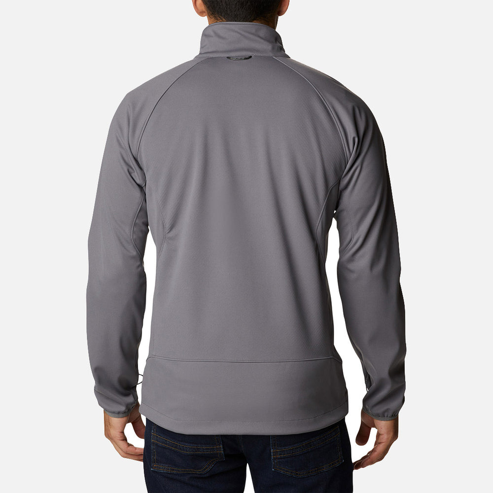 נראה אדם מאחור, לבוש במעיל Softshell של ColumbiaIL מדגם Canyon Meadows בצבע אפור בהיר, עם שרוולים ארוכים וצווארון גבוה, בשילוב עם ג'ינס כחול כהה, כשהוא עומד על רקע לבן חלק.