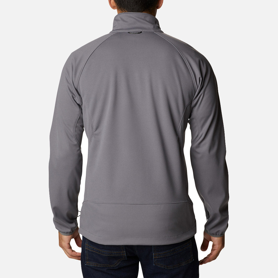 נראה אדם מאחור, לבוש במעיל Softshell של ColumbiaIL מדגם Canyon Meadows בצבע אפור בהיר, עם שרוולים ארוכים וצווארון גבוה, בשילוב עם ג'ינס כחול כהה, כשהוא עומד על רקע לבן חלק.