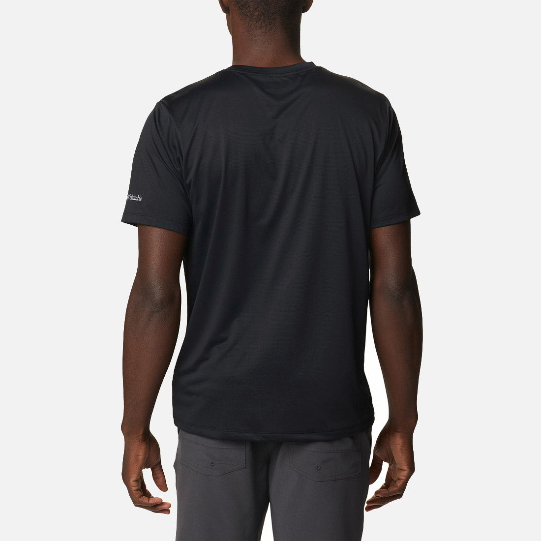 אדם עומד עם גבו למצלמה, לבוש בחולצת Columbia Hike Graphic S/S T מבית columbiaIL ובמכנסיים אפורים כהים, על רקע לבן חלק.