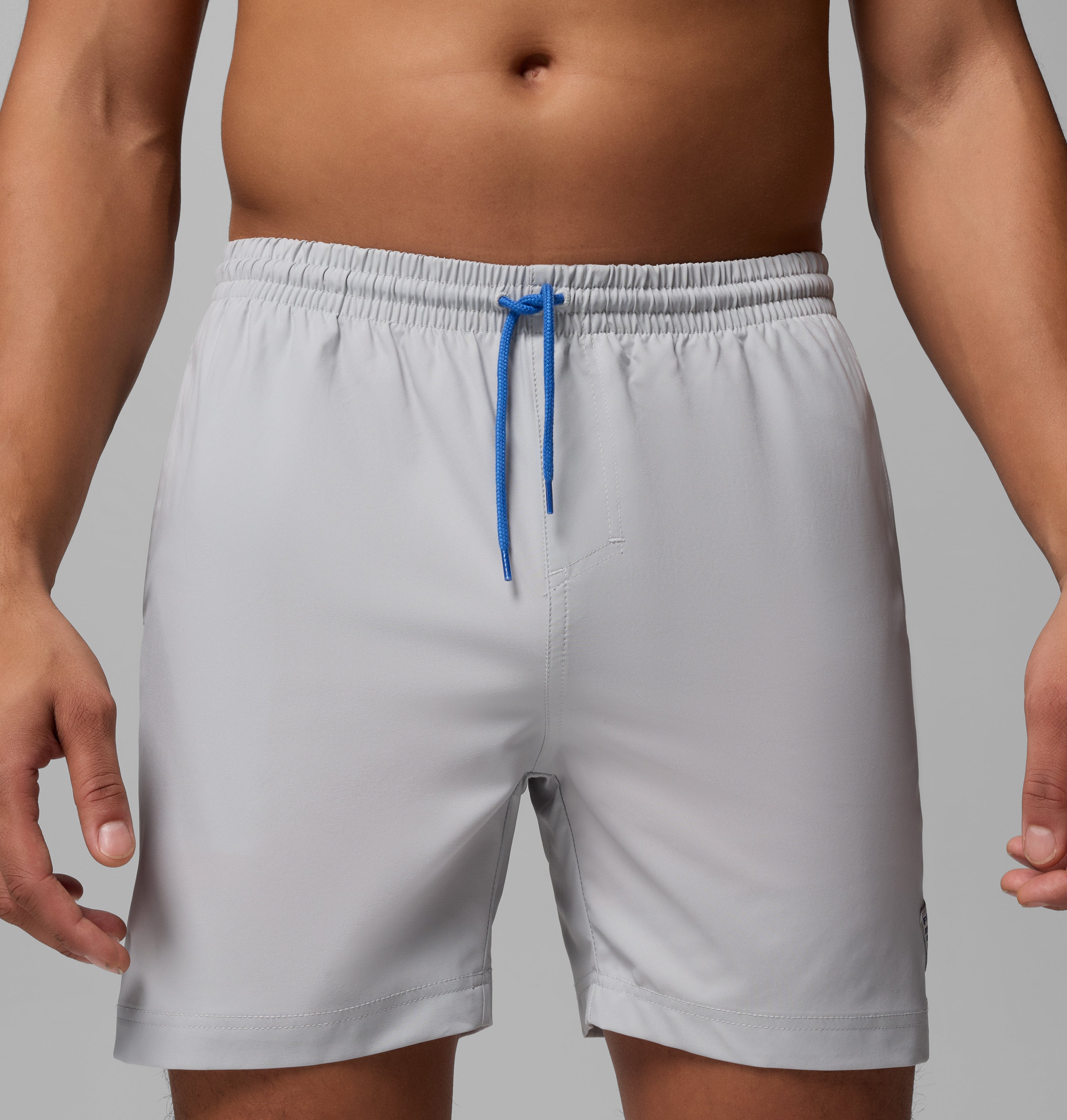 אדם מדגמן את מכנסי השחייה PFG Rambler Water Short של קולומביה ישראל בצבע אפור בהיר עם שרוך כחול, כשהוא עומד על רקע אפור אחיד. נראים רק פלג גופו העליון ורגליו העליונות.