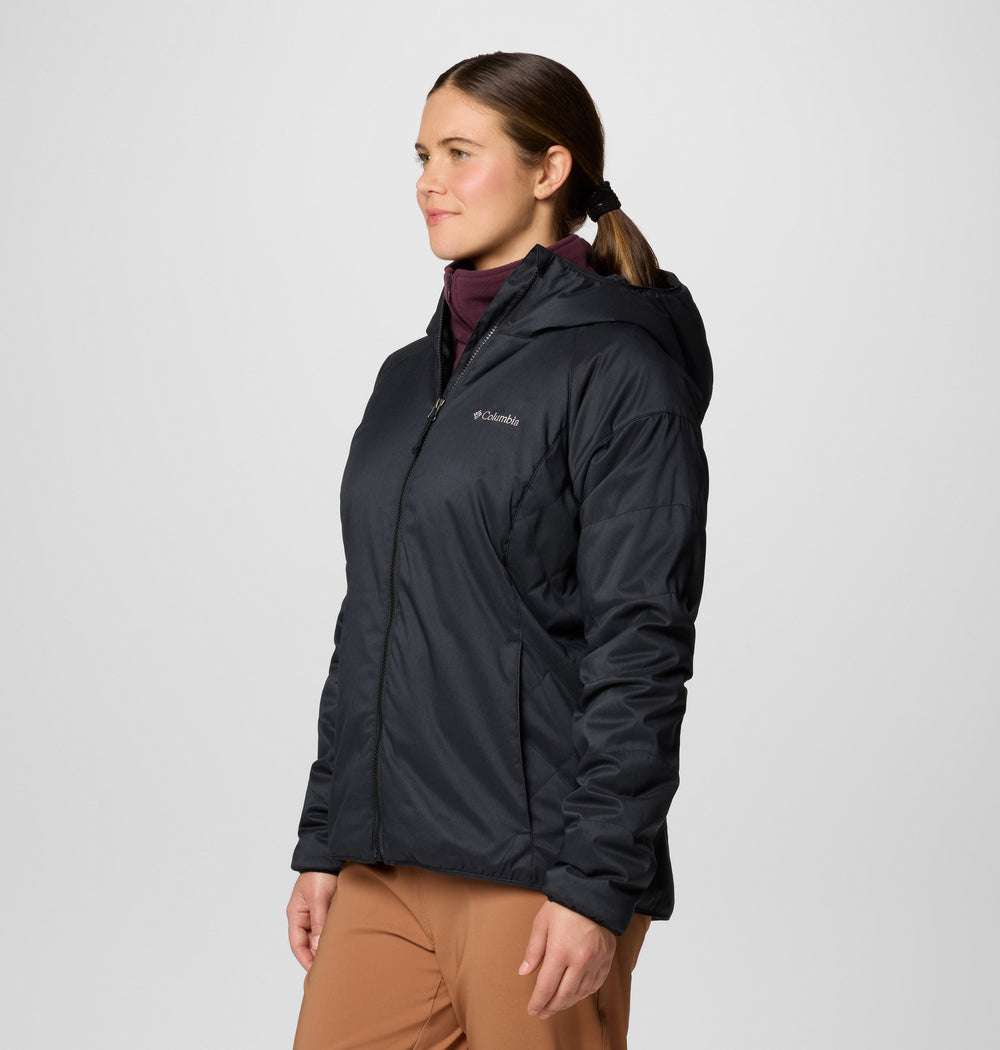 אישה עם שיער חום אסוף לקוקו לובשת מעיל שחור מדגם "ColumbiaIL Kruser Ridge III Plush Sfts Jkt" מעל חולצה בצבע בורדו ומכנסיים בצבע חום בהיר, ועומדת על רקע אפור בהיר חלק.