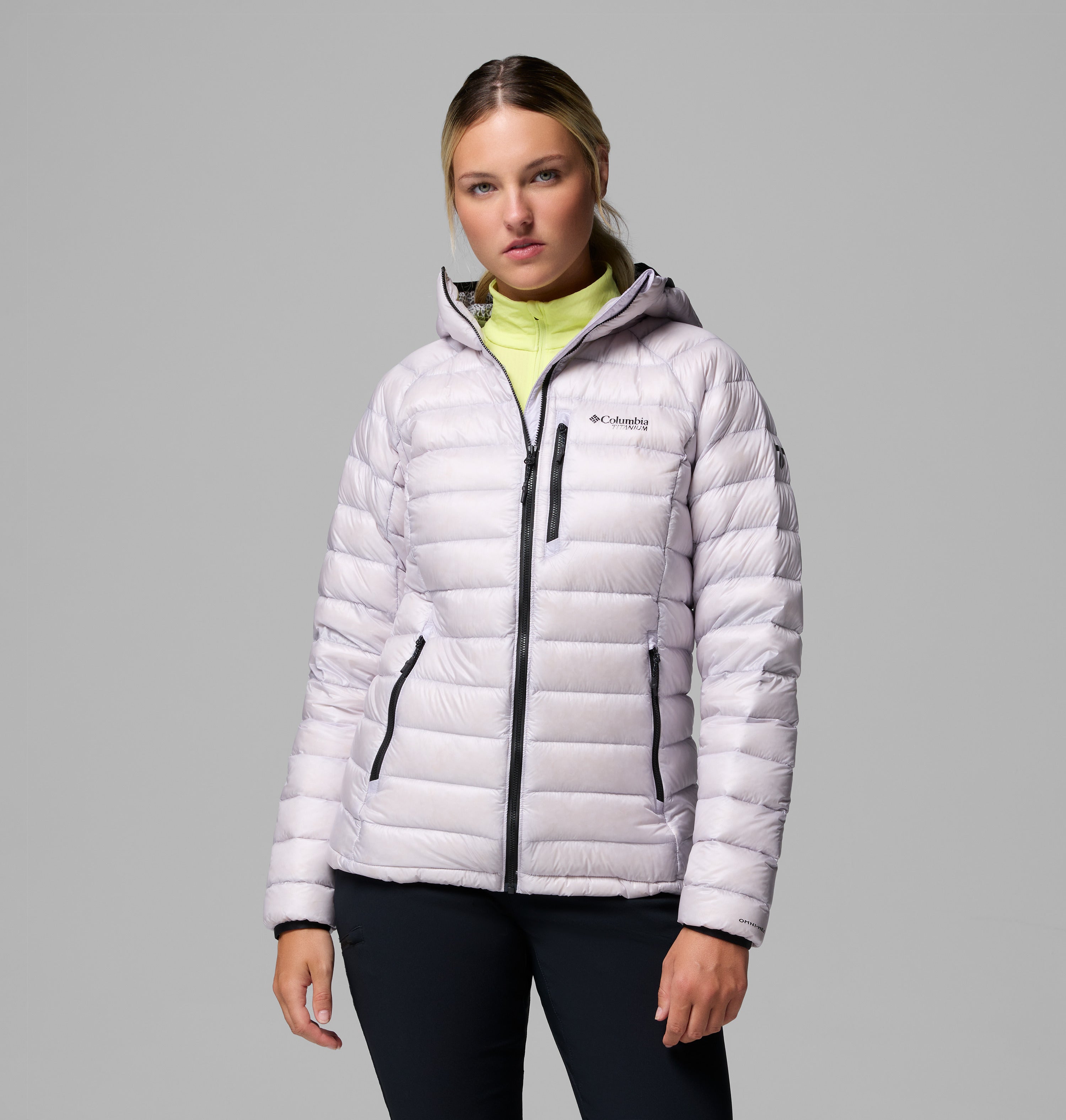 אישה לבושה במעיל פוך עם ברדס מדגם Arctic Crest של המותג ColumbiaIL, עם רוכסנים שחורים, עומדת על רקע אפור אחיד ומביטה במצלמה במבט ניטרלי.