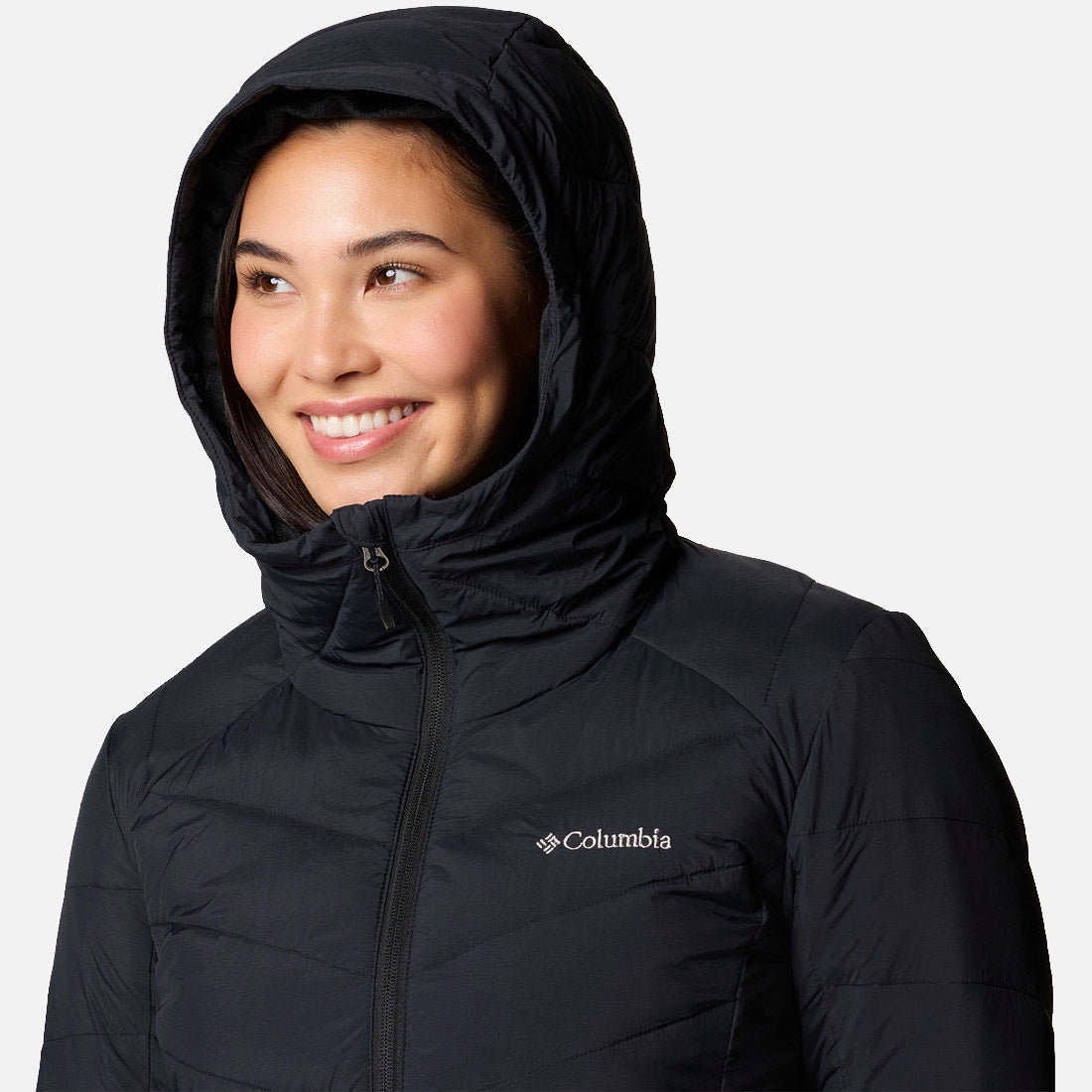 אדם הלובש מעיל "קולומביה IL Joy Peak II Mid" בצבע שחור מחייך ומביט מעט הצידה על רקע בהיר וחד-גוני.