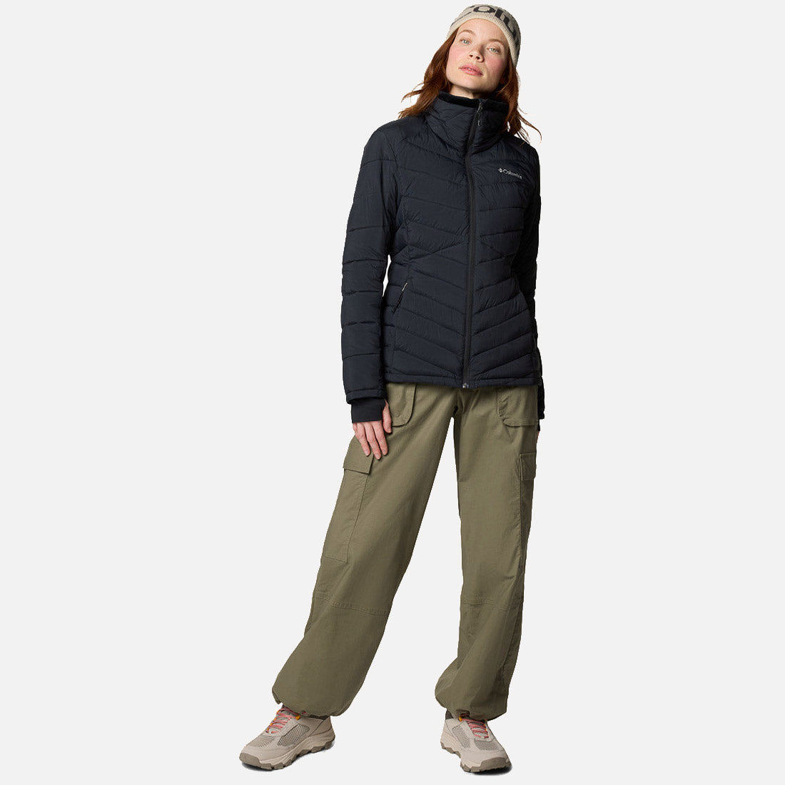 אישה לבושה במעיל שחור מדגם "Joy Peak II Full Zip" של המותג "קולומביה" (קולומביה, אילינוי), מכנסי קרגו בצבע ירוק זית, נעלי ספורט בצבע בז' וסרט לראש, עומדת על רקע לבן חלק, מביטה כלפי מעלה בתנוחה נינוחה.