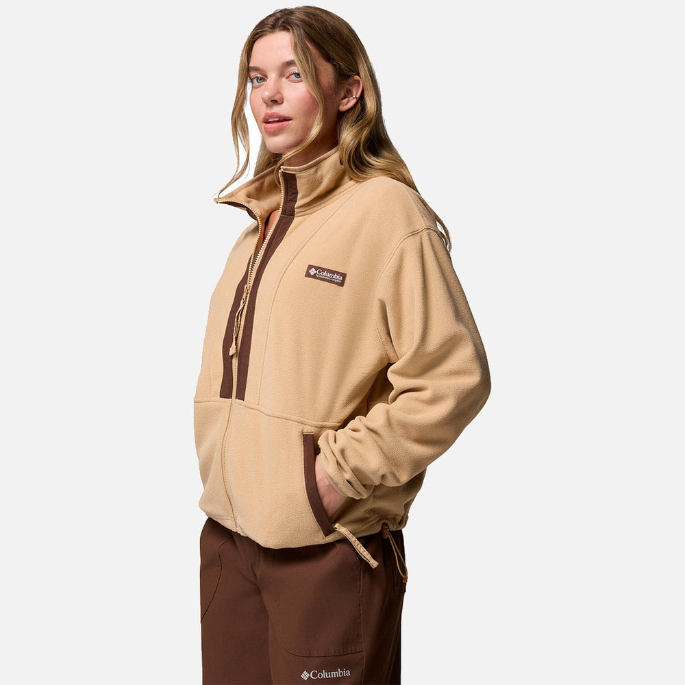אישה לבושה במעיל "Backbowl II Full Zip W" בצבע בז' של המותג ColumbiaIL ובמכנסיים חומים, ידיה בכיסים, עומדת על רקע לבן — בגדי חוץ מסוגננים לכל עונה.