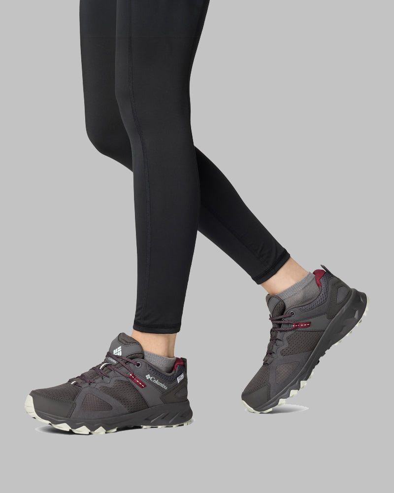 אדם הלובש טייץ שחור ונעלי קולומביה ישראל Peakfreak Hera Outdry עם נגיעות אדומות עומד על רקע אפור אחיד, כשנראים רק רגליו וכפות רגליו.