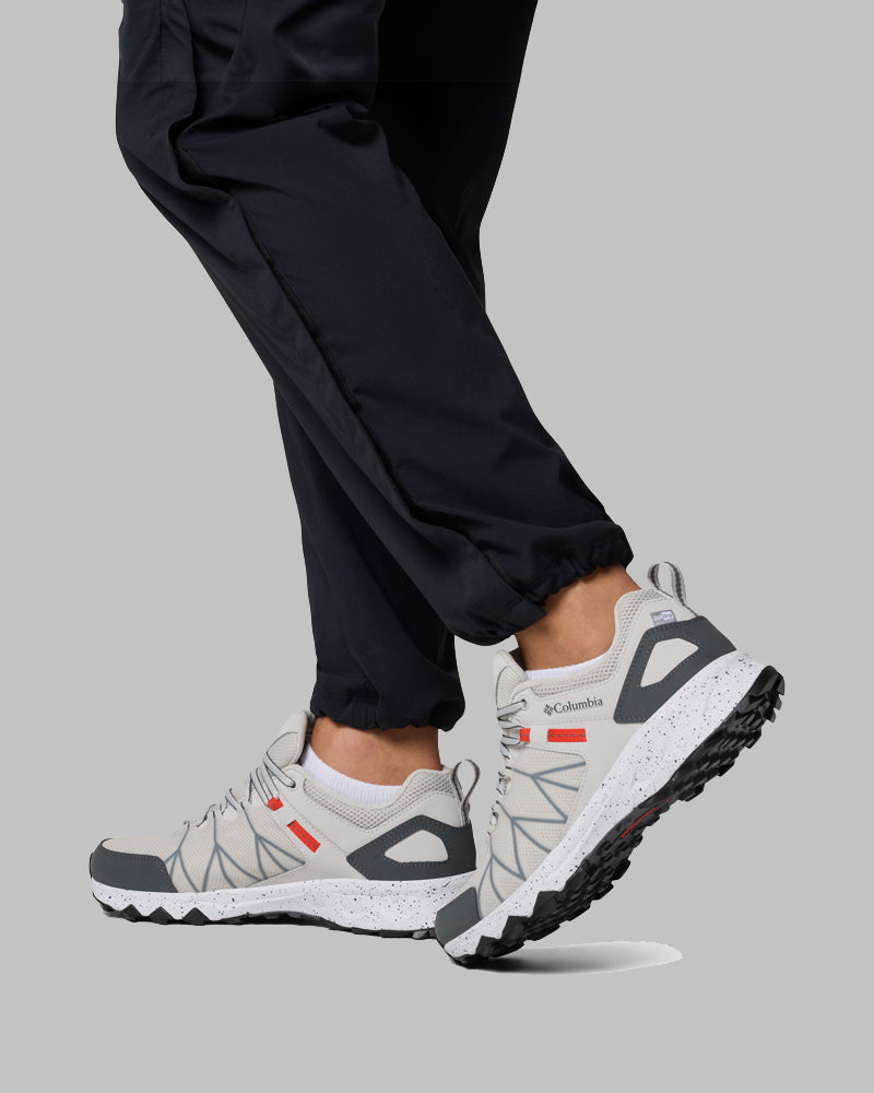אדם לבוש במכנסיים שחורים ונעלי ספורט מסוג Columbia IL Peakfreak II Outdry בצבע אפור בהיר עם נגיעות אדומות צועד קדימה על רקע אפור אחיד; הנעליים מתאפיינות בעיצוב שטח מחוספס ובסוליות שחורות וחזקות.