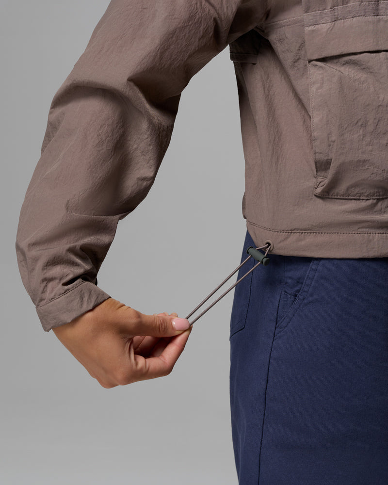 אדם מהדק את שרוך התחתון של מעיל רוח קצר מסוג "Paracutie Cropped Cargo" של קולומביה ישראל בצבע חום בהיר, כשהוא לובש מכנסי קרגו כחולים. התמונה מדגישה את מותניו בזמן שהוא מתאים את המעיל.