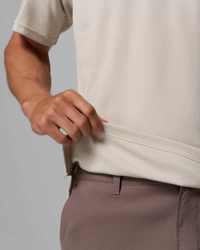 אדם הלובש חולצת קולומביה ישראל Alpine Chill Pro Short Sleeve Crew בצבעי בז' וסגול בהיר, ומכנסיים, אוחז בשולי החולצה ביד אחת ומרים אותם קלות.