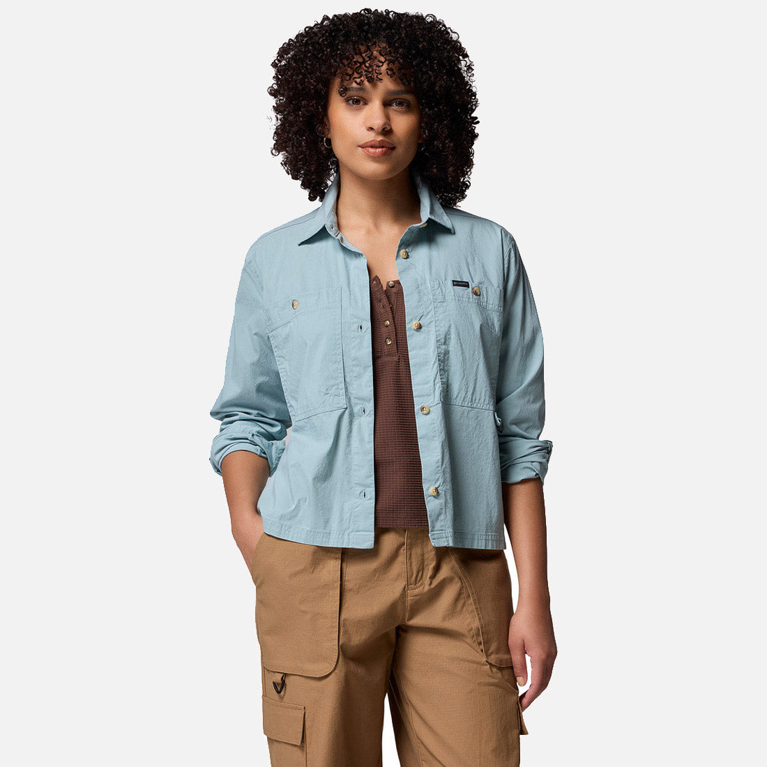 אישה עם שיער מתולתל לובשת את חולצת "Brea Falls Woven Long Sleeve Shirt" של קולומביה ישראל בצבע תכלת, מעל חולצה חומה ומכנסי קרגו בצבע חום בהיר, ועומדת כשידה האחת בכיס על רקע לבן חלק.