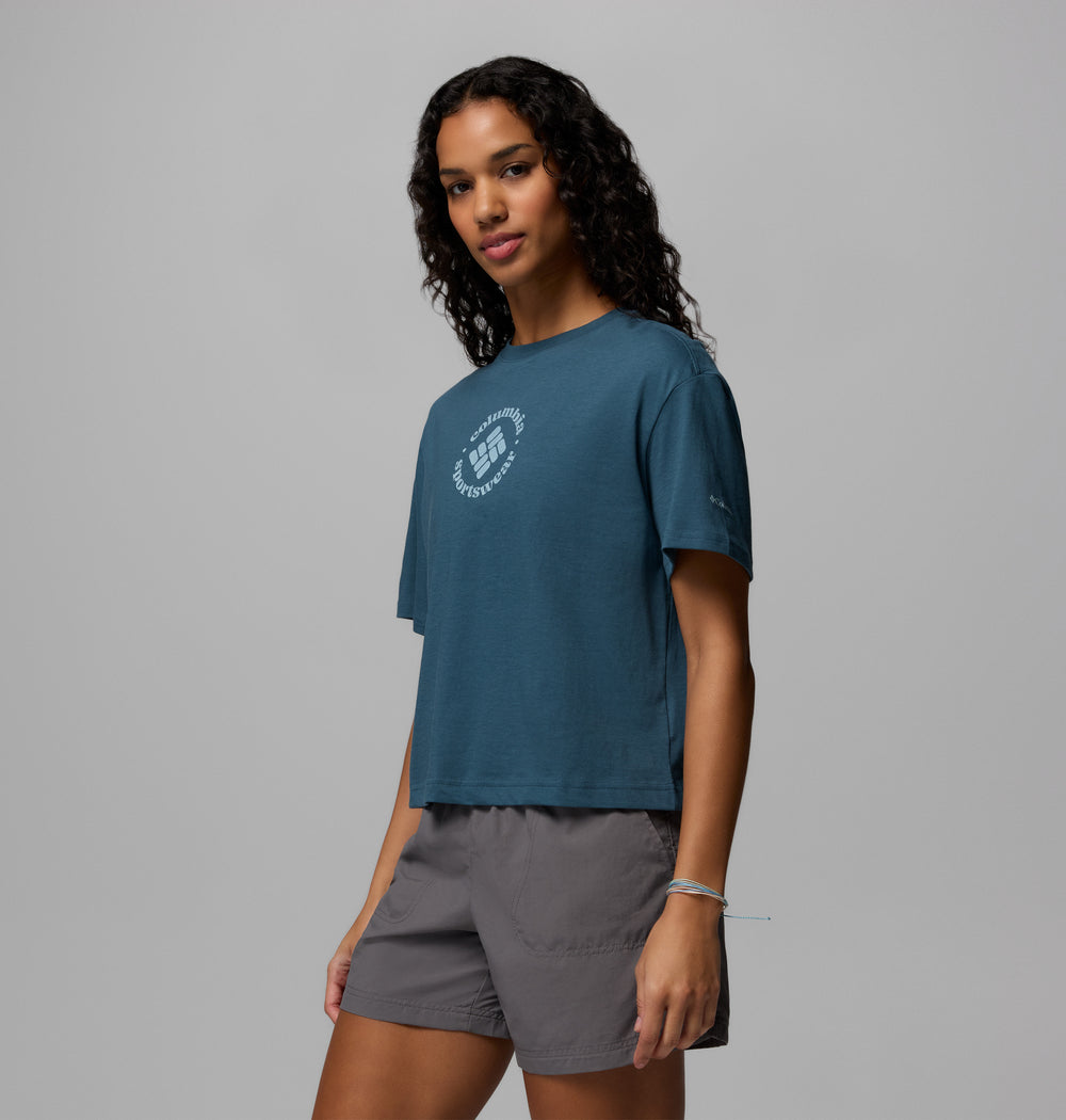 אישה עם שיער ארוך ומתולתל מדגמנת את חולצת ה-Graphic Relaxed Crop Short Sleeve Shirt של קולומביה ישראל בצבע טורקיז, בשילוב עם מכנסיים קצרים אפורים. היא עומדת על רקע אפור בהיר, מביטה לצד עם הבעה נינוחה.