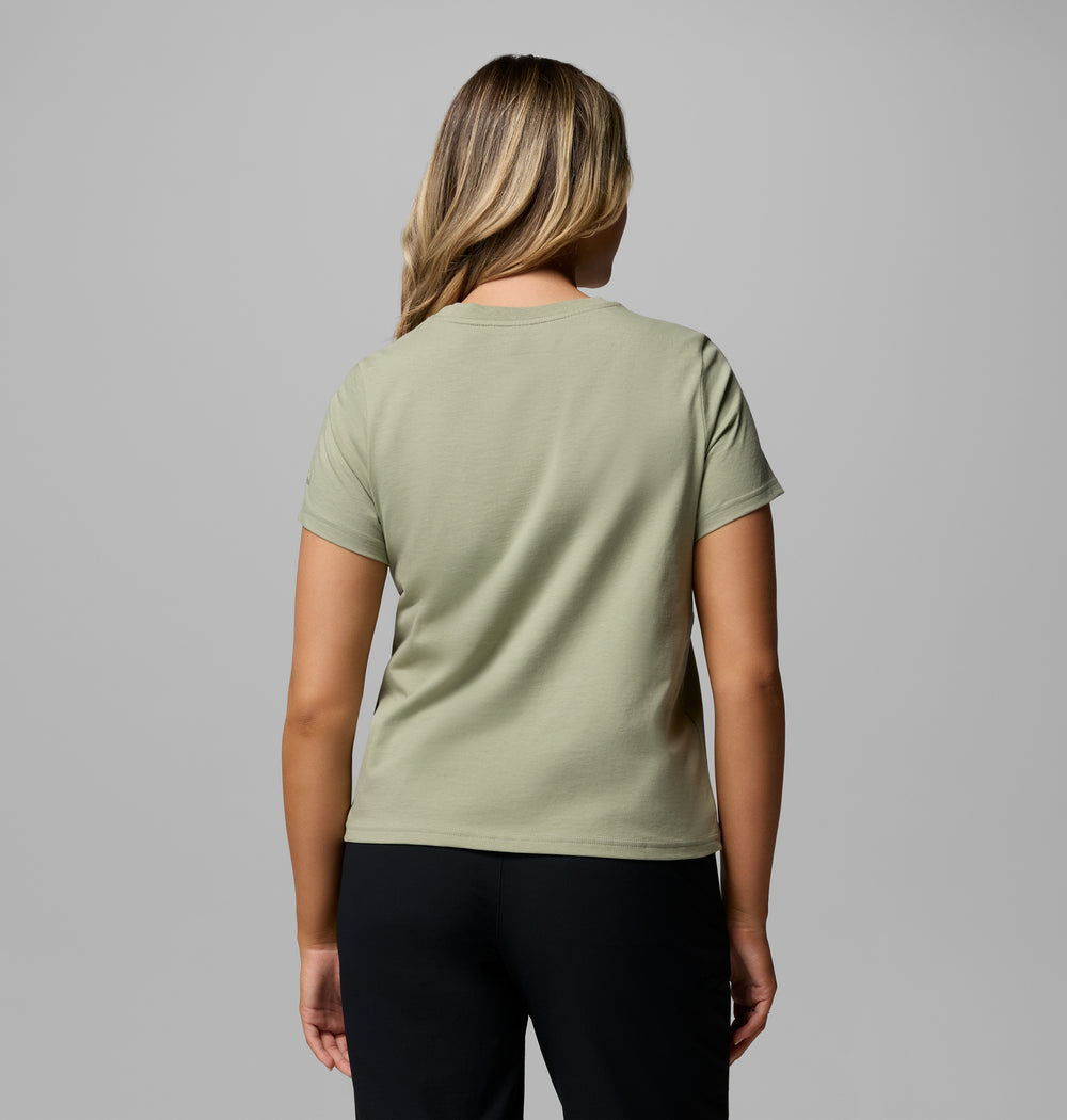 אישה בעלת שיער חום בהיר מדגמנת את חולצת ה-Rolling Bend Graphic Short Sleeve Shirt של קולומביה ישראל בצבע ירוק בהיר ומכנסיים שחורים, כשהיא עומדת עם גבה למצלמה על רקע אפור אחיד.