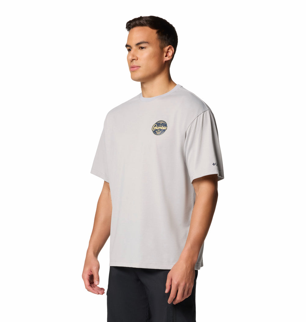 גבר לבוש בחולצת טי-שירט "Cedar Trail Graphic T" של קולומביהIL בצבע אפור בהיר, עם הדפס מעגלי קטן על החזה השמאלי, ומכנסיים קצרים כהים, עומד על רקע לבן חלק.