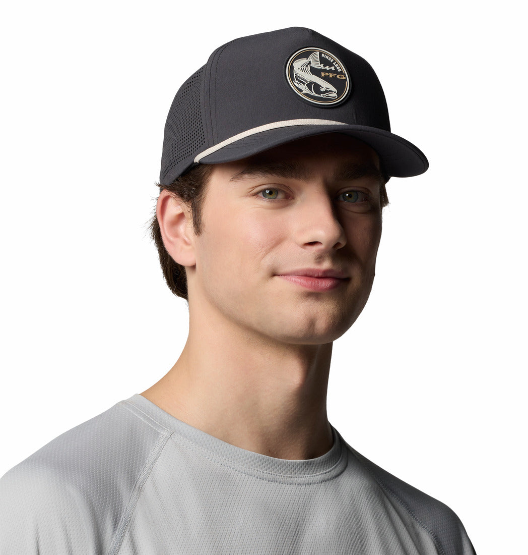 צעיר חובש כובע "Tidal Pursuit 3D Stretch Snap Back" בצבע כהה של המותג ColumbiaIL, עם לוגו של דג, ולובש חולצה אפורה בהירה, מחייך קלות על רקע לבן חלק.