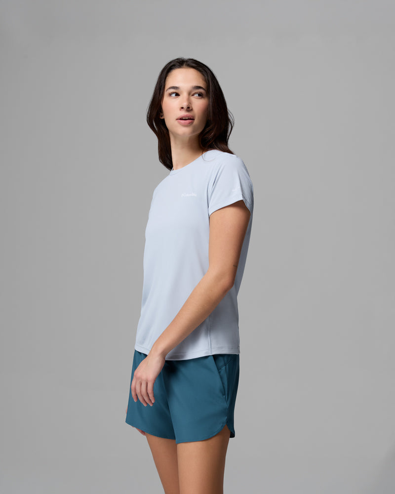 אישה עם שיער חום ארוך לובשת את חולצת ה-Zero Rules Light Short Sleeve Crew של קולומביה ישראל בצבע תכלת, בשילוב עם מכנסיים קצרים בצבע טורקיז, עומדת על רקע אפור אחיד ומביטה מעט הצידה.