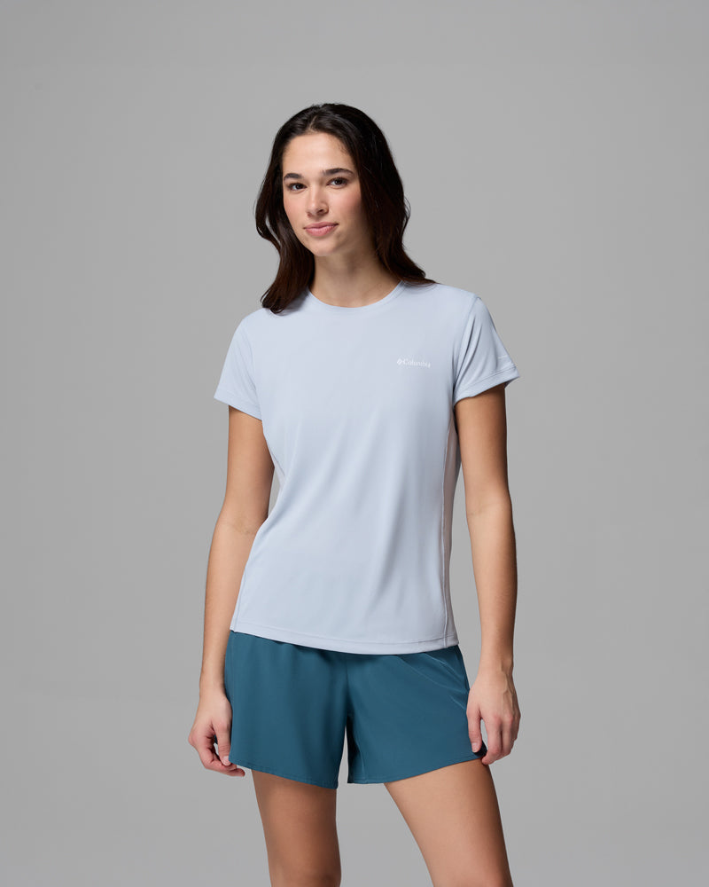 אישה עם שיער חום ארוך לובשת את חולצת ה-Zero Rules Light Short Sleeve Crew של קולומביה ישראל בצבע תכלת ומכנסיים קצרים בצבע טורקיז, ועומדת על רקע אפור אחיד. היא פונה קדימה עם חיוך קל, ומדגימה את סגנון ה-ZERO RULES הטבעי.