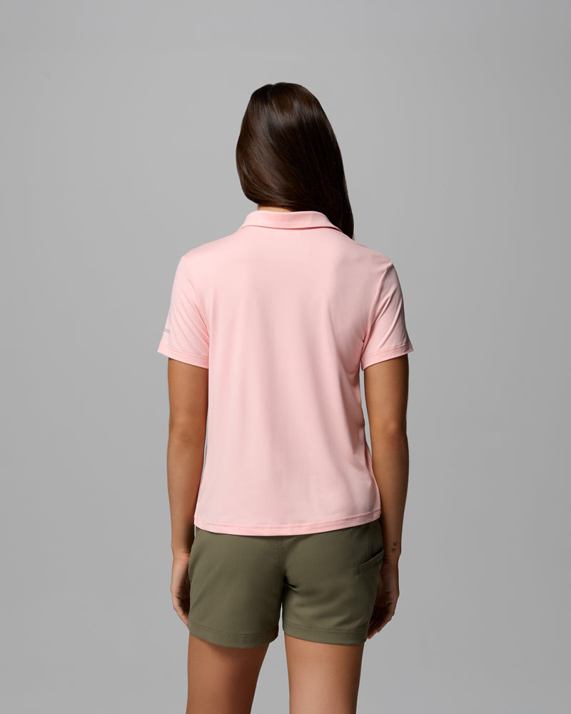A woman with long brown hair, seen from behind, wears a light pink Parsons Point Polo W and olive green shorts against a plain gray background.