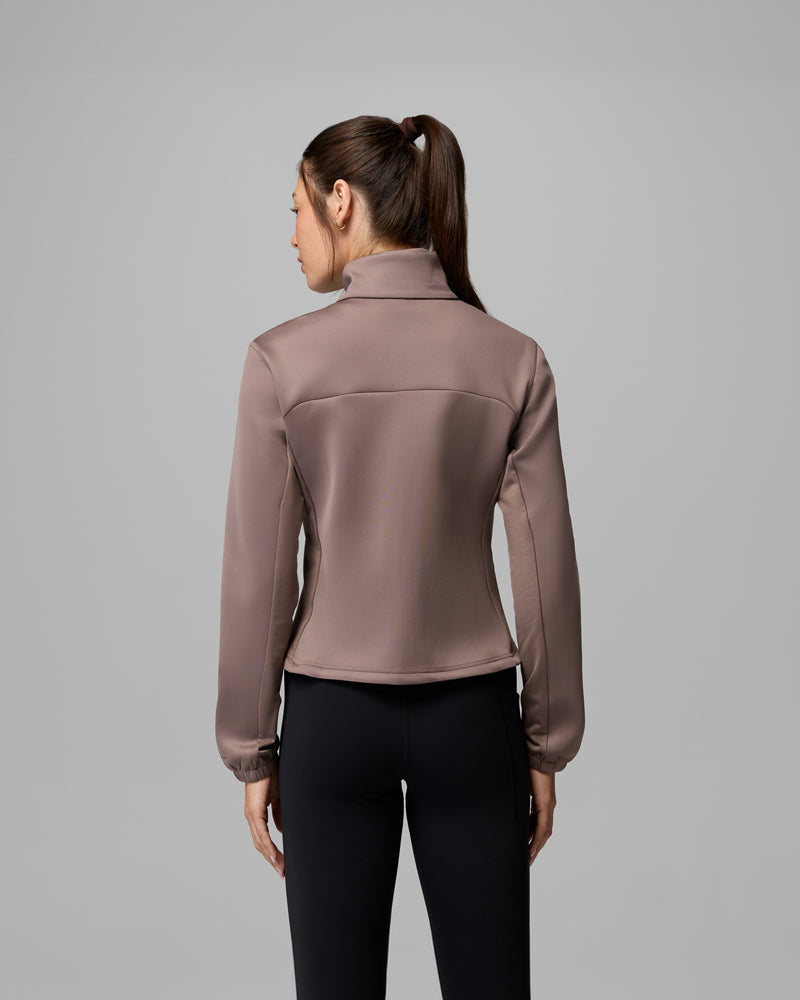 נראית אישה עם קוקו חום, מצולמת מאחור, לבושה בז'קט בצבע טופ של קולומביה ישראל מדגם "Sloan Crest Spacer Fleece Full Zip" ומכנסיים צמודים שחורים, עומדת על רקע אפור אחיד.