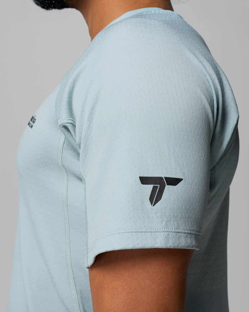 תמונת תקריב מהצד של חולצת ה-Driventure Short Sleeve Shirt של קולומביה ישראל, חולצת טי ספורטיבית לגברים בצבע תכלת עם שרוולים קצרים ולוגו גיאומטרי שחור על השרוול. פניו של הדוגמן אינן נראות.