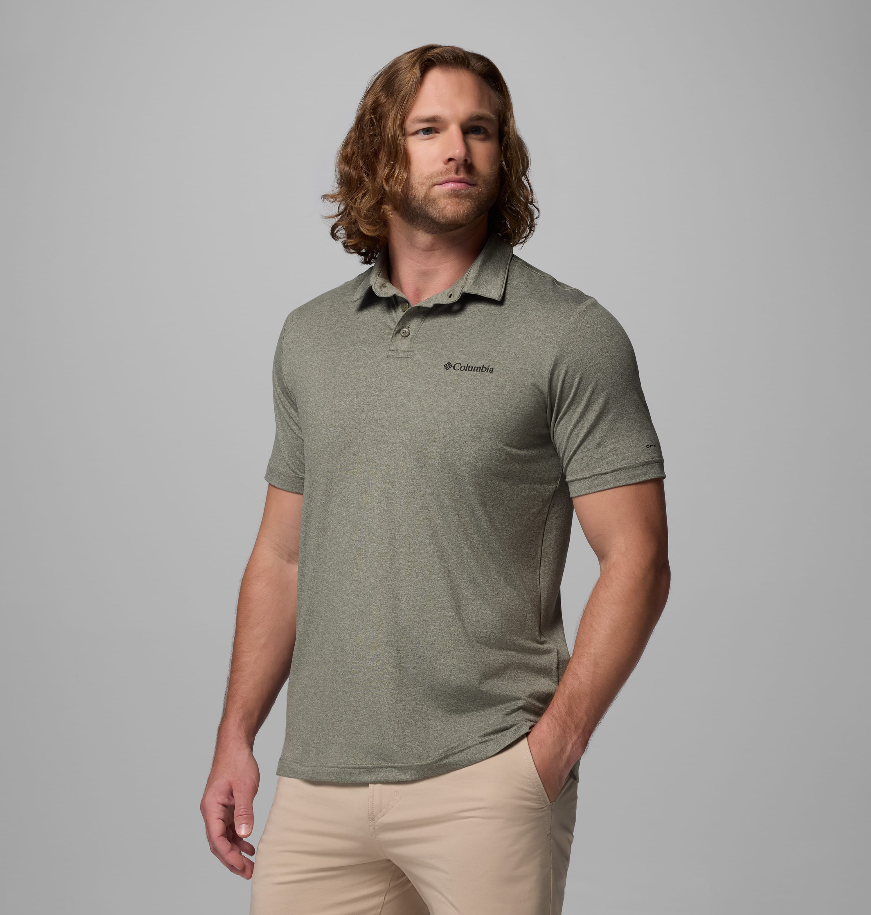 גבר בעל שיער ארוך וגלי לובש חולצת פולו "Parsons Point Polo M" בצבע אפור בהיר של קולומביה ישראל ומכנסיים בצבע בז', עומד כש יד אחת בכיסו על רקע אפור אחיד.