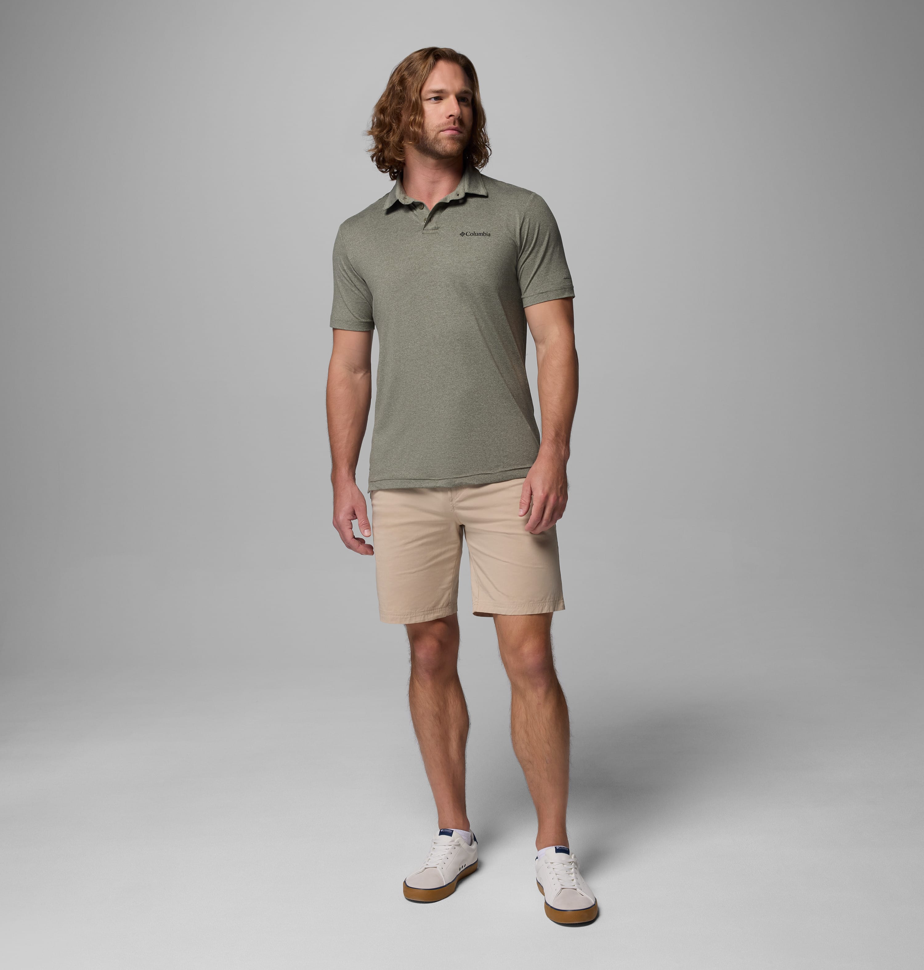 גבר בעל שיער ארוך לובש חולצת פולו "Parsons Point Polo M" בצבע זית עם שרוולים קצרים של קולומביה ישראל, מכנסיים קצרים בצבע בז' ונעלי ספורט לבנות, והוא עומד על רקע אפור בהיר אחיד.