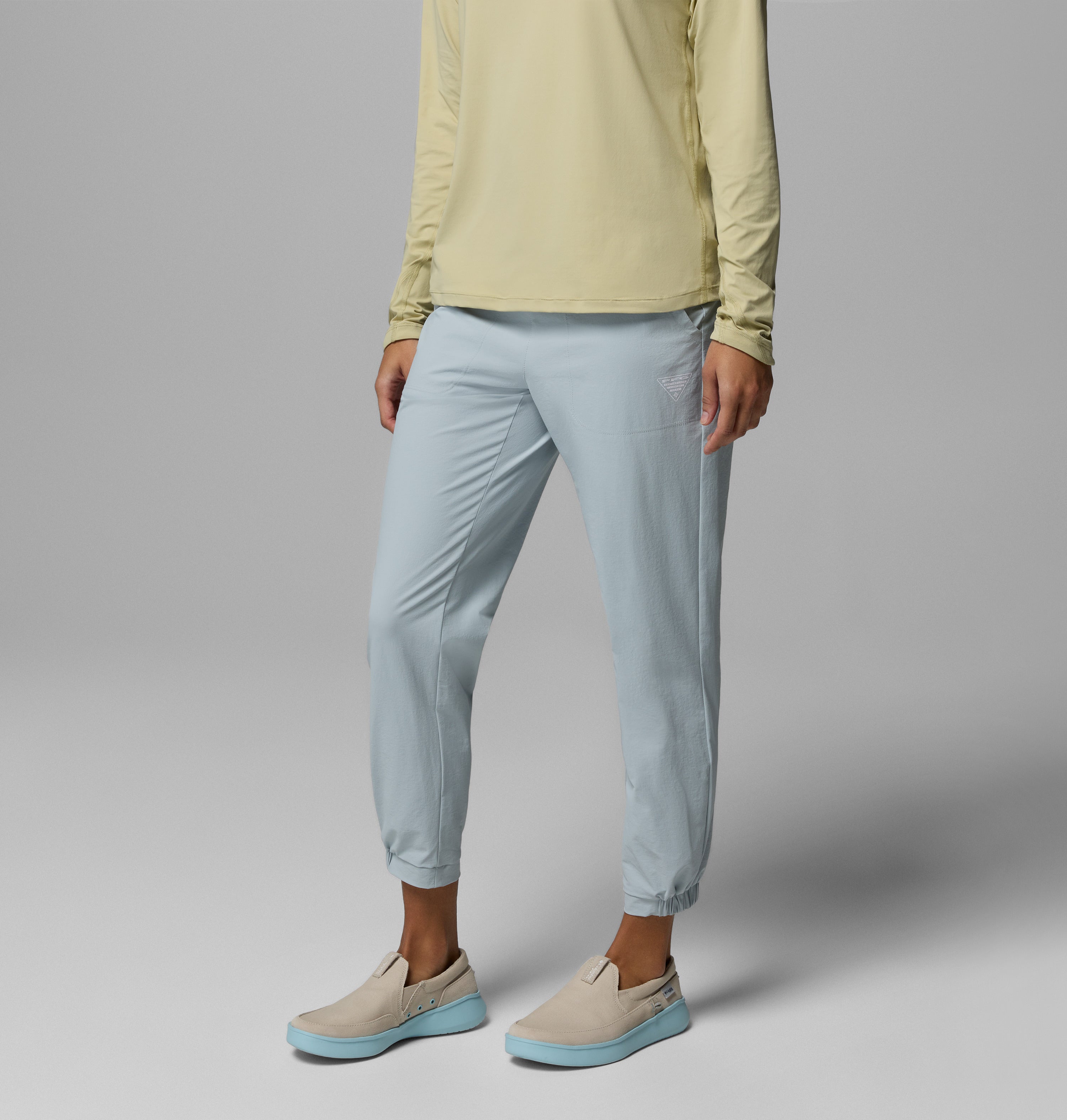 אדם עומד על רקע אפור אחיד, לבוש במכנסי Wild Cast Pant של קולומביה ישראל בצבע תכלת, בשילוב עם חולצה ארוכת שרוולים בצבע צהוב בהיר ונעלי סליפ-און בצבע בז' עם סוליות תכלת. ראשו אינו נראה.