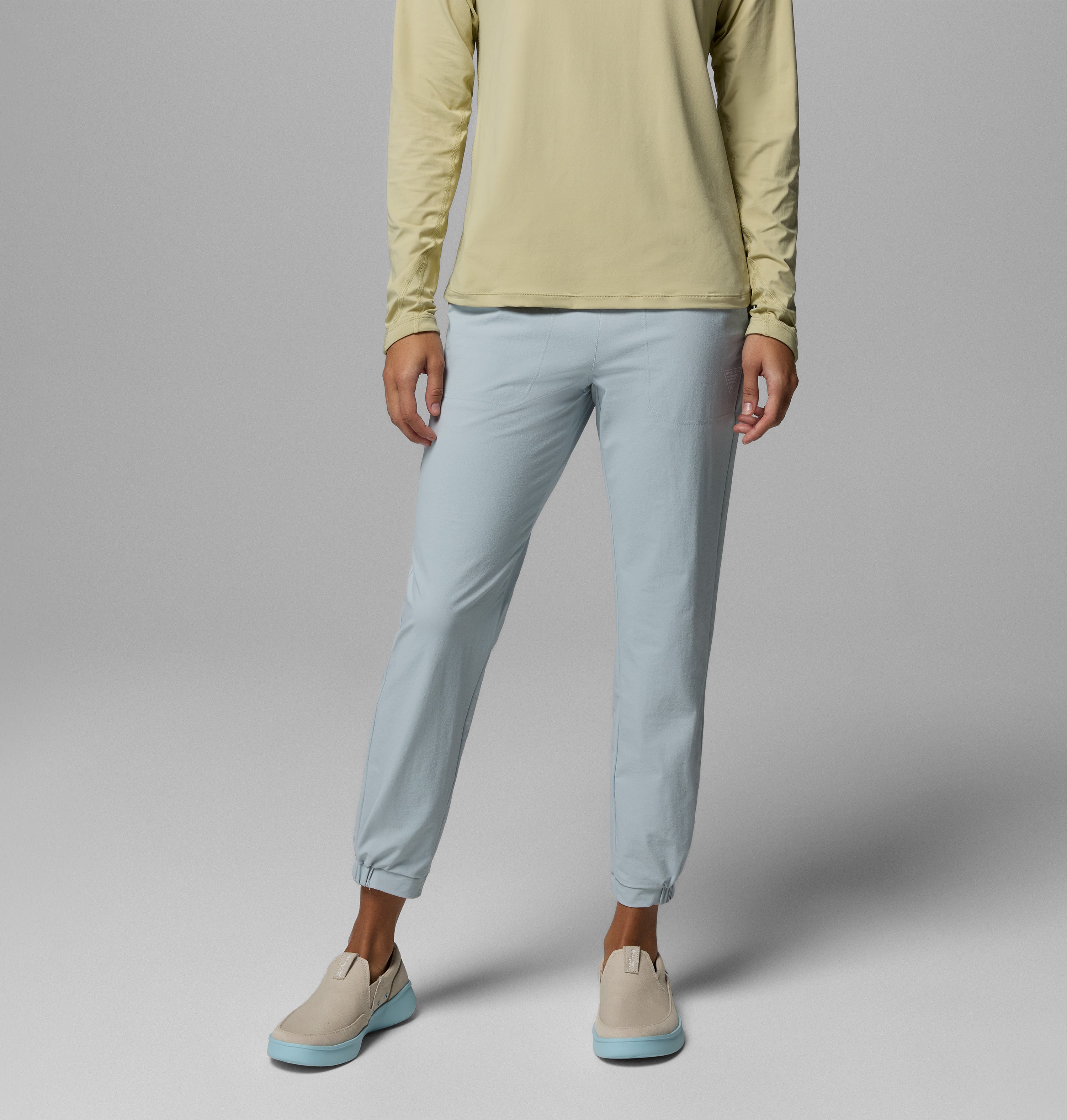 אדם עומד על רקע אפור, לבוש במכנסי Wild Cast Pant של קולומביה ישראל בצבע תכלת ובחולצה ארוכת שרוולים בצבע צהוב בהיר. נעלי סליפ-און בצבע בז' עם סוליות תכלת משלימות את המראה. פניו אינם נראים.