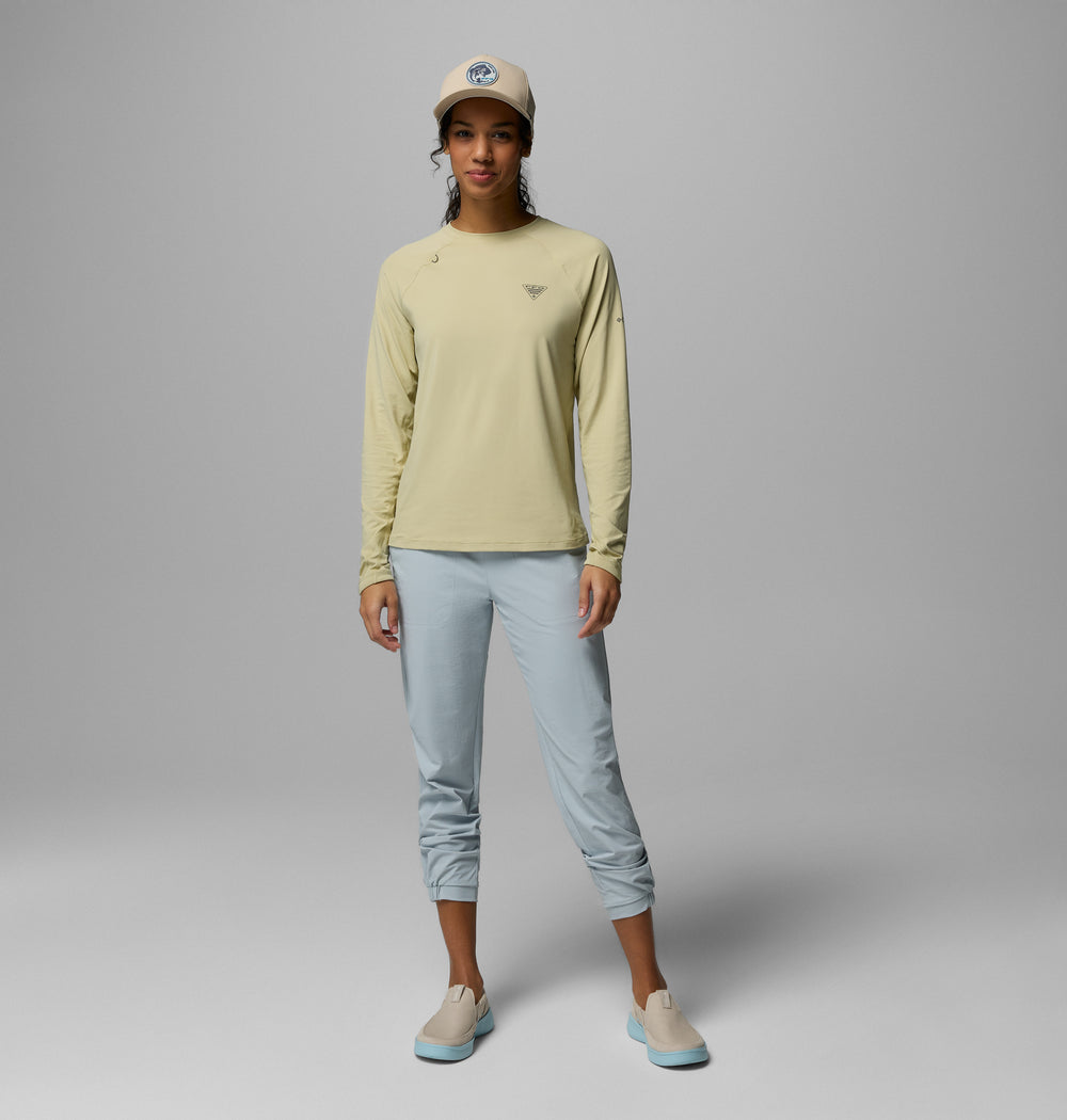 אישה עומדת בסטודיו, לבושה במכנסי "Wild Cast Pant" של קולומביה ישראל בצבע תכלת, חולצה בצבע בז' עם שרוולים ארוכים, נעליים ללא שרוכים בצבע בז' עם סוליות כחולות וכובע בייסבול בצבע בז', על רקע אפור בהיר אחיד.