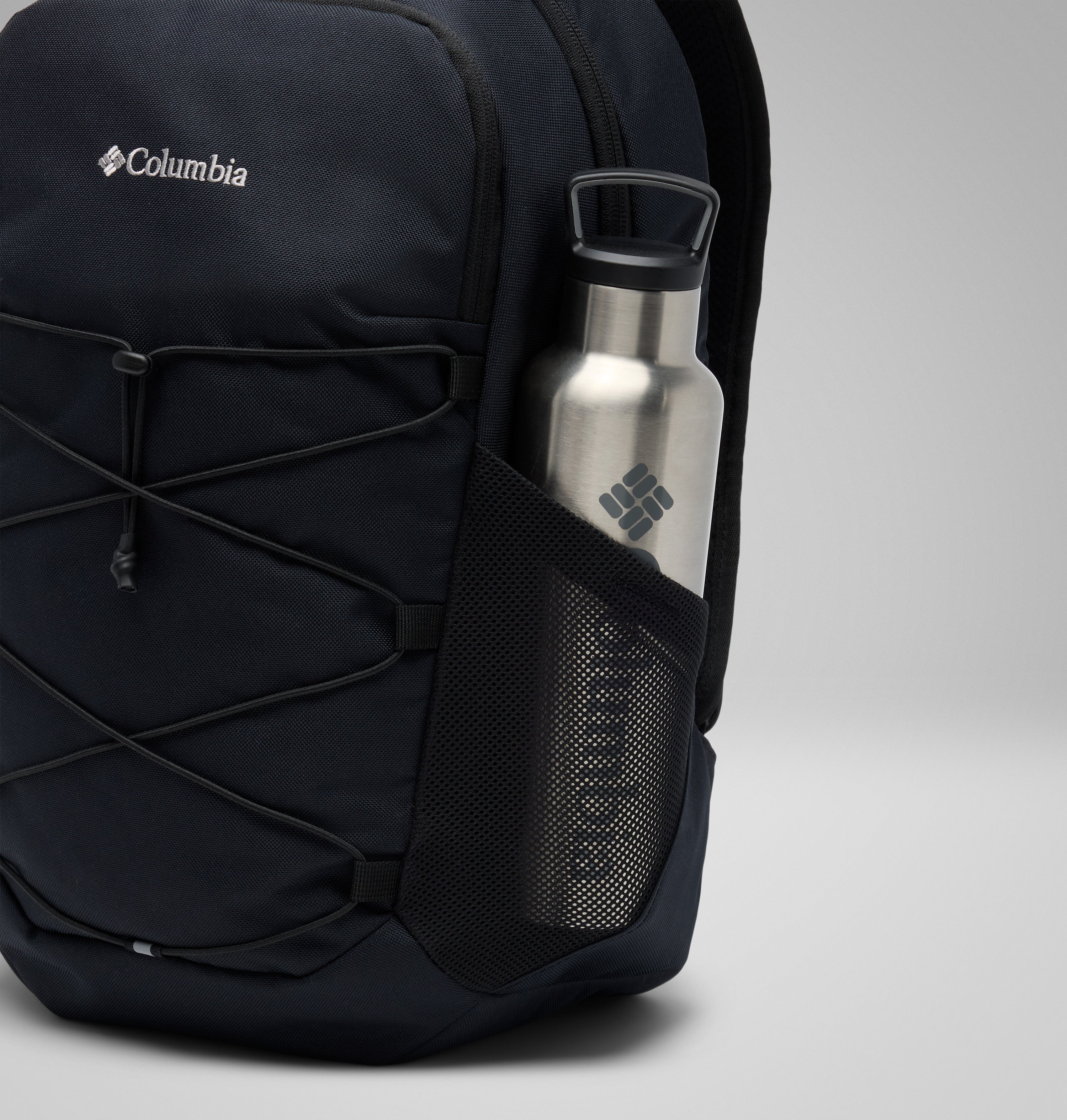 תיק גב שחור מדגם "Atlas Explorer 20L" של קולומביה ישראל, עם חוט אלסטי בחזית וכיס צדדי מרשת המכיל בקבוק מים מפלדת אל-חלד, שניהם נושאים את הלוגו של קולומביה ישראל, על רקע אפור בהיר אחיד.