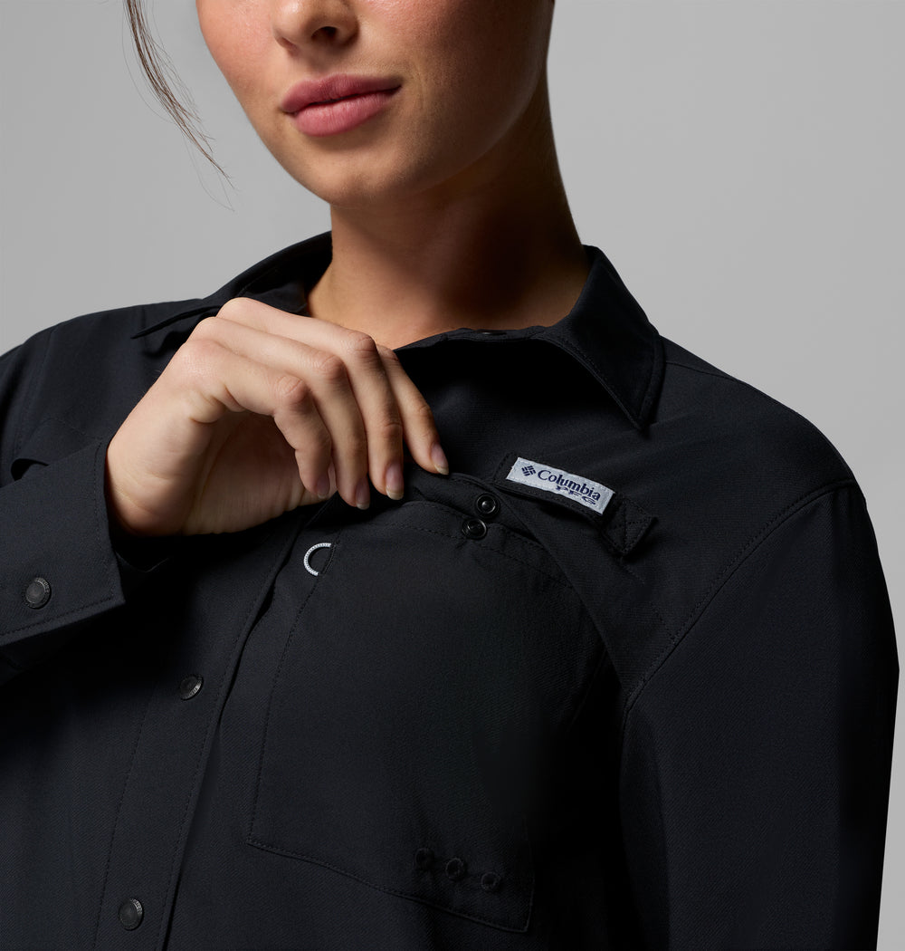 אדם לבוש בחולצת קולומביה ישראל PFG Castback Woven L/S שחורה פותח את הכפתורים של כיס החזה כדי לחשוף את תווית הלוגו, כאשר רק החלק התחתון של פניו נראה על רקע אפור אחיד.