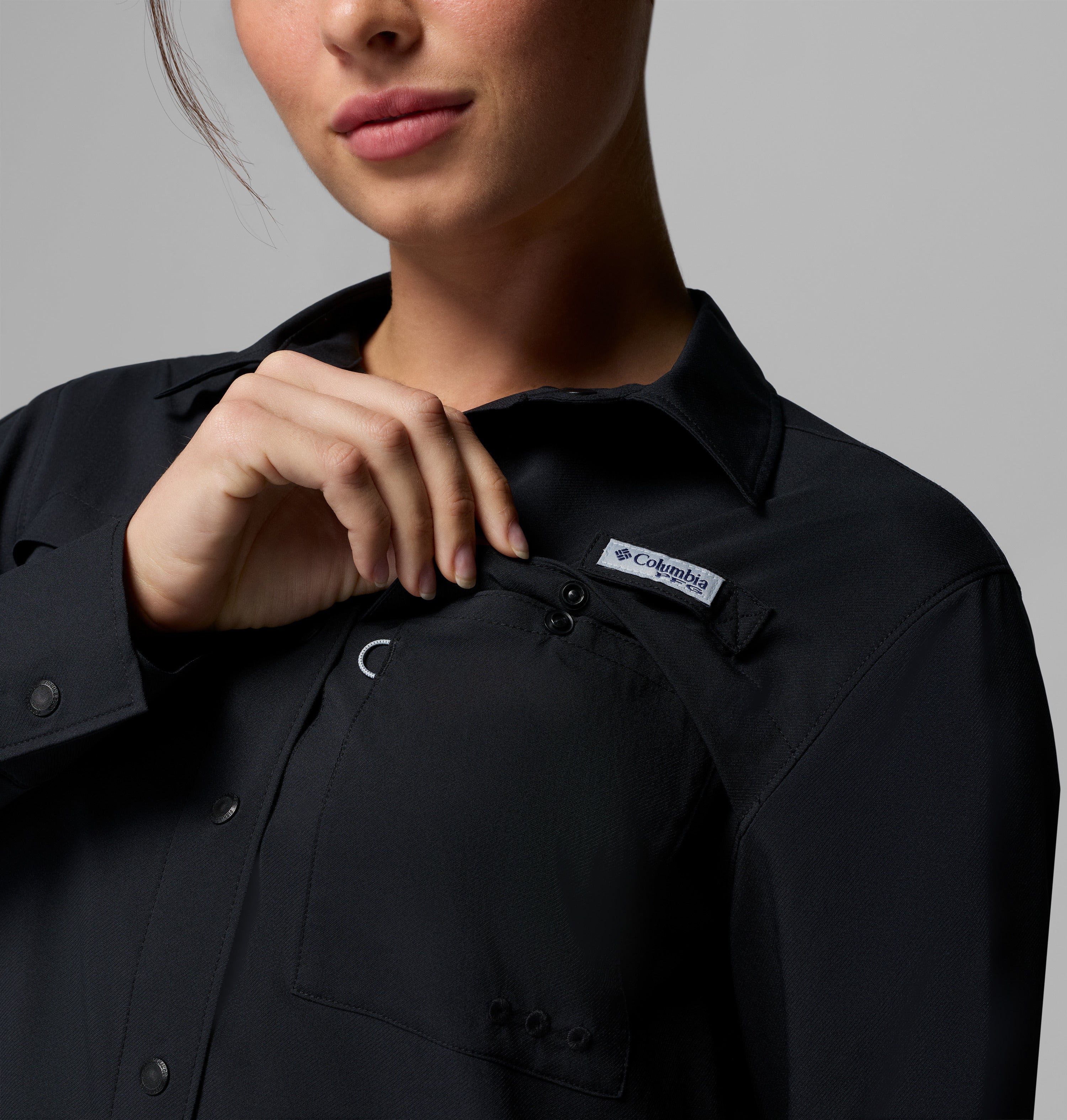 אדם לבוש בחולצת קולומביה ישראל PFG Castback Woven L/S שחורה פותח את הכפתורים של כיס החזה כדי לחשוף את תווית הלוגו, כאשר רק החלק התחתון של פניו נראה על רקע אפור אחיד.
