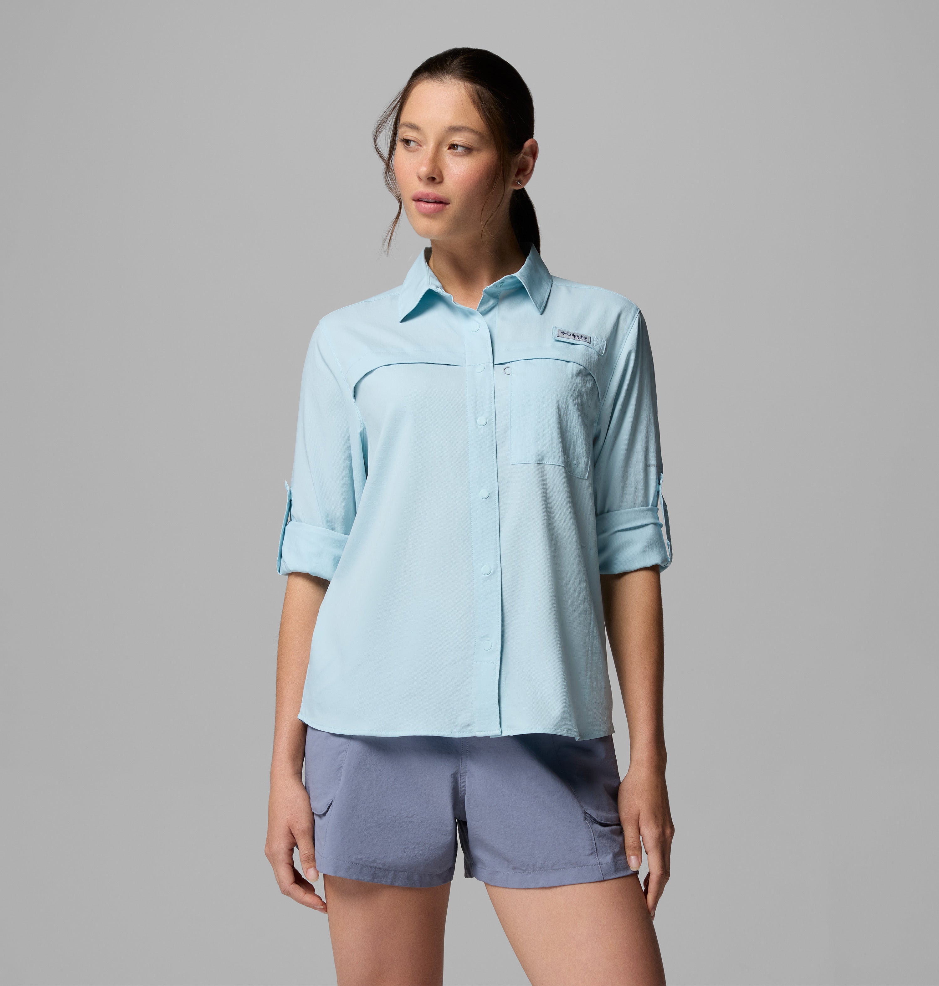 אישה לבושה בחולצת קולומביה ישראל PFG Castback Woven L/S בצבע תכלת, עם שרוולים מופשלים, ומכנסיים קצרים אפורים, עומדת על רקע אפור אחיד ומביטה מעט הצידה.