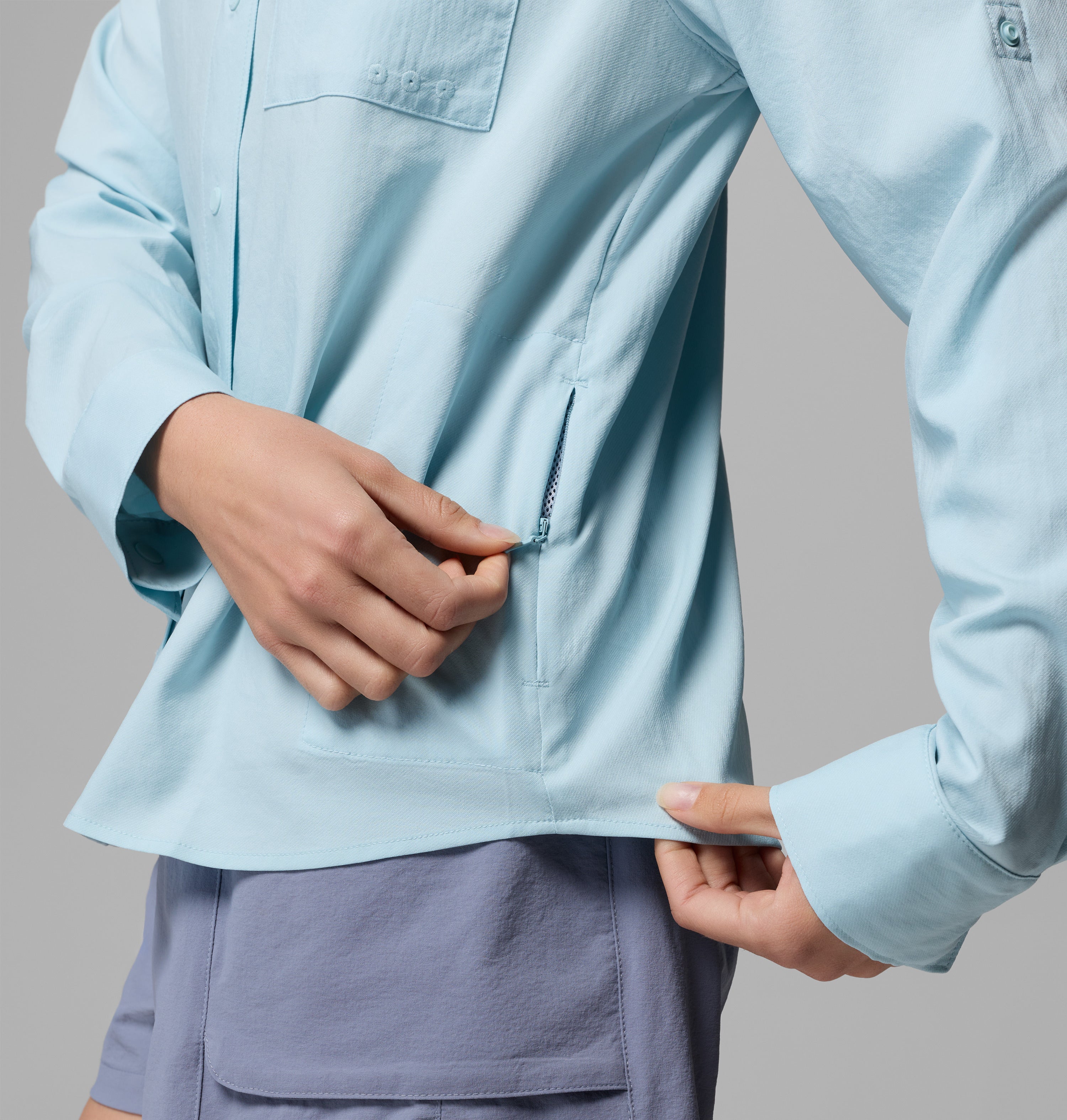אדם לבוש בחולצת קולומביה ישראל PFG Castback Woven L/S בצבע תכלת ובמכנסיים קצרים בצבע לבנדר פותח את רוכסן הכיס הצדדי בחולצה, על רקע אפור אחיד.