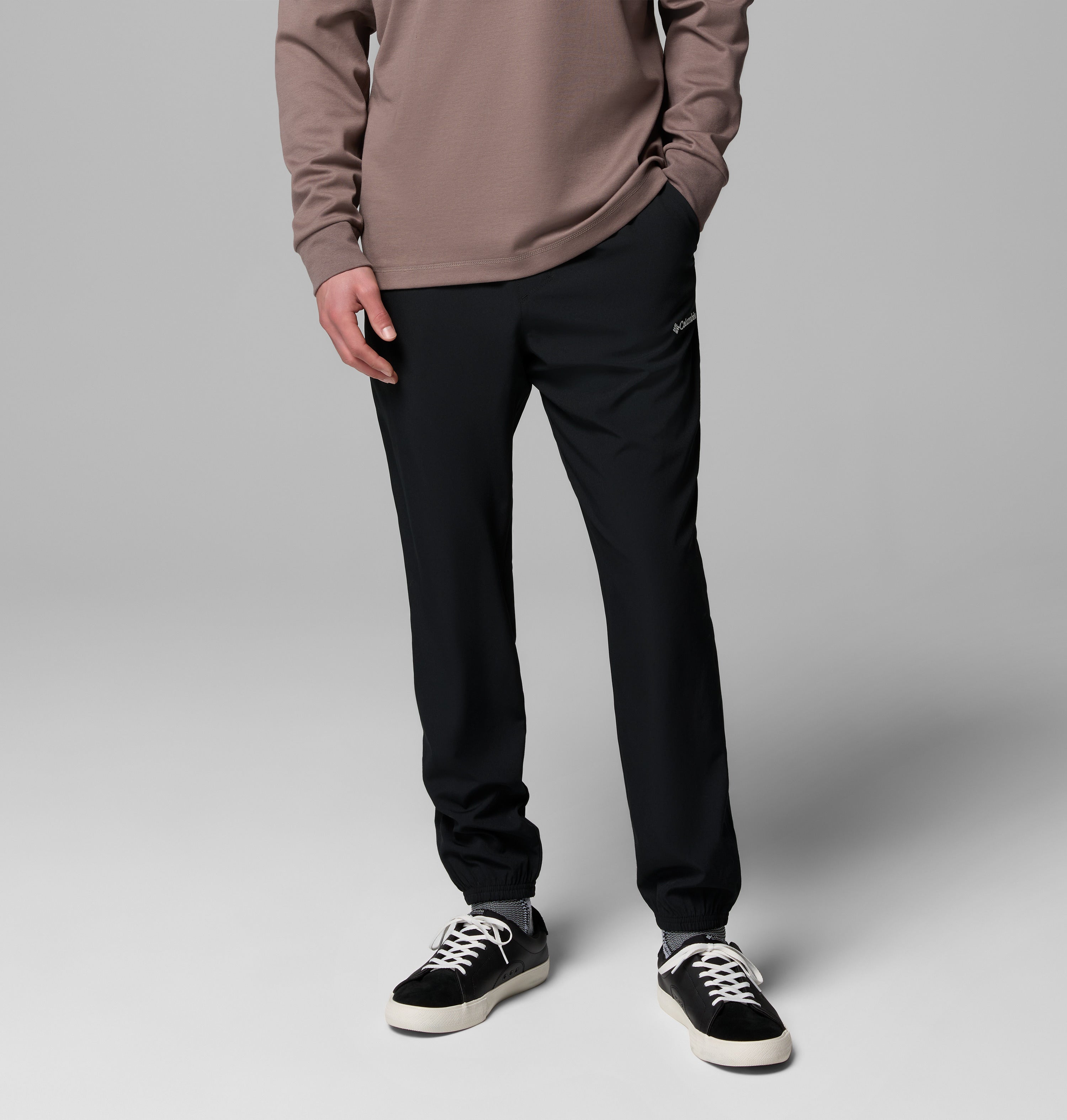 אדם לבוש במכנסי ריצה מדגם "Vital Valley Jogger" של קולומביה ישראל, חולצה בצבע טופ עם שרוולים ארוכים, ונעלי ספורט בשחור-לבן, עומד כשכף יד אחת בכיס על רקע אפור אחיד.