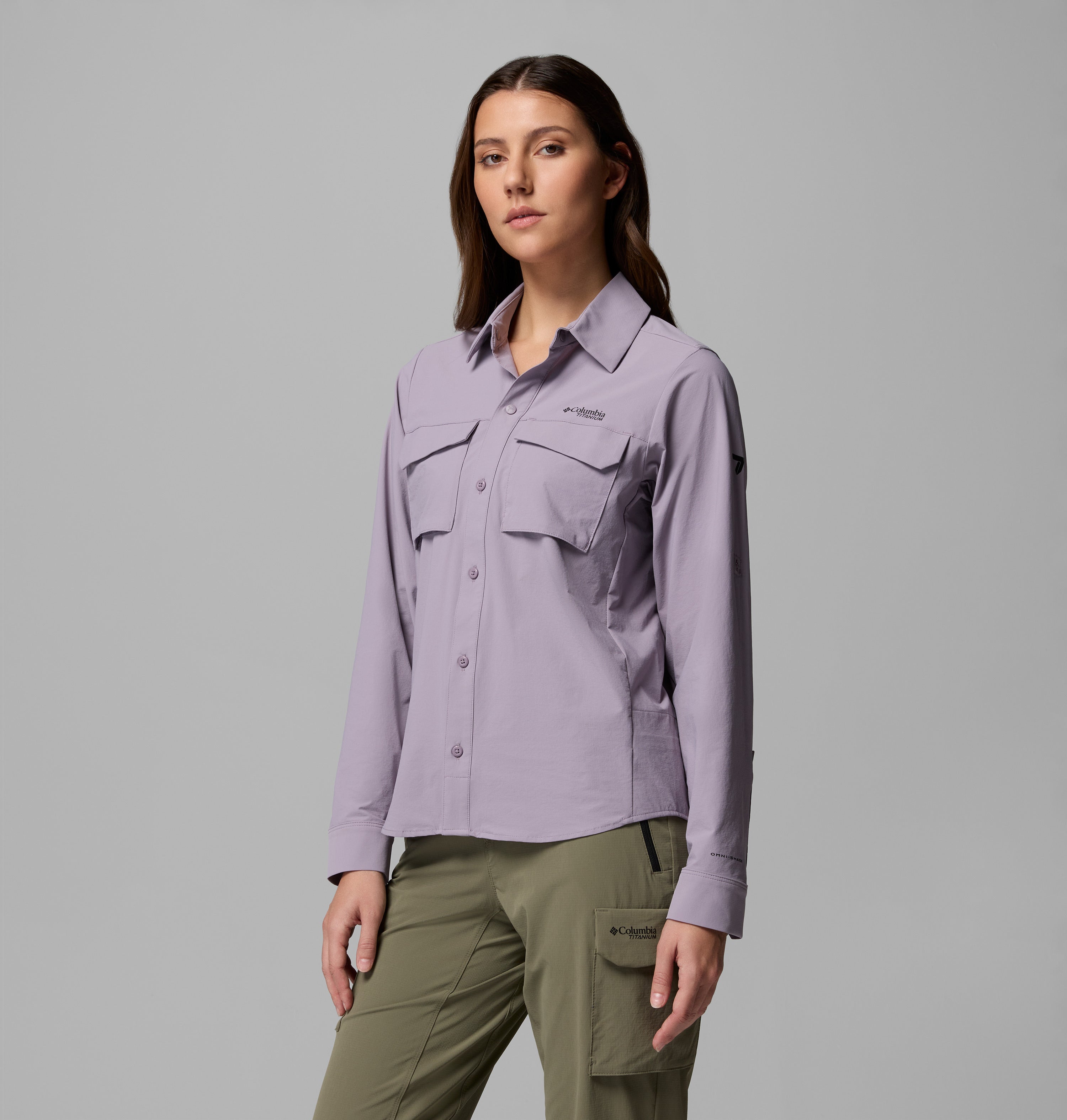 אישה עומדת פונה קדימה, לבושה בחולצת "Silver Ridge Elite Woven L/S" בצבע סגול בהיר מבית קולומביה ישראל, עם שני כיסים קדמיים, ומכנסי קרגו בצבע ירוק זית, כשהיא מצטלמת על רקע אפור אחיד.