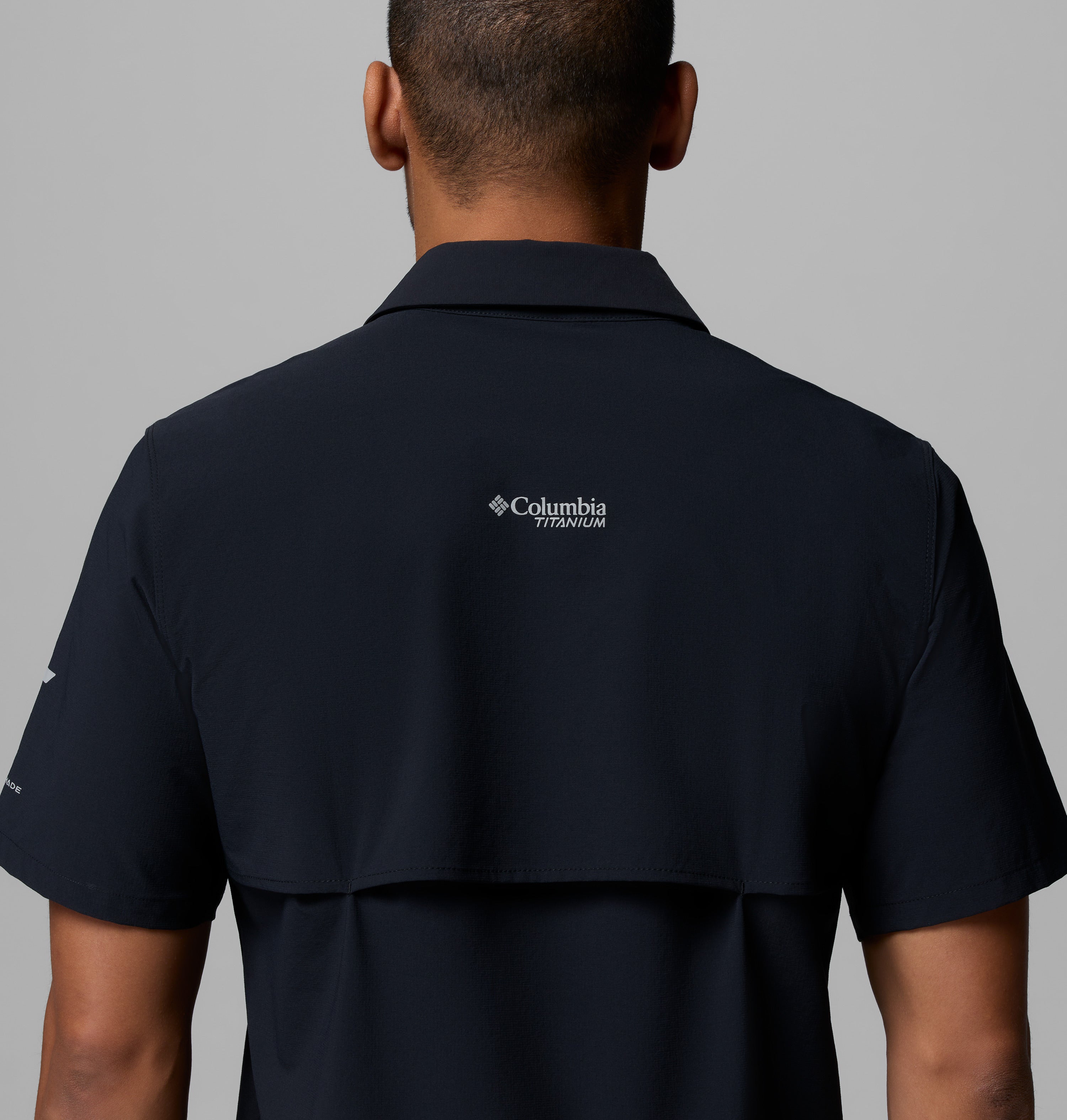 נראית דמות מאחור, לבושה בחולצת "Silver Ridge Elite S/S Woven" בצבע כהה מבית קולומביה ישראל, על רקע אפור אחיד. לוגו המותג מופיע בחלק העליון של הגב בחולצה הארוגה והמסוגננת הזו.
