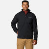 גבר עומד עם הפנים קדימה, לבוש במעיל Softshell שחור של ColumbiaIL Ascender מעל חולצה בורדו ומכנסיים בצבע חום בהיר, יד אחת בכיס, על רקע לבן חלק.